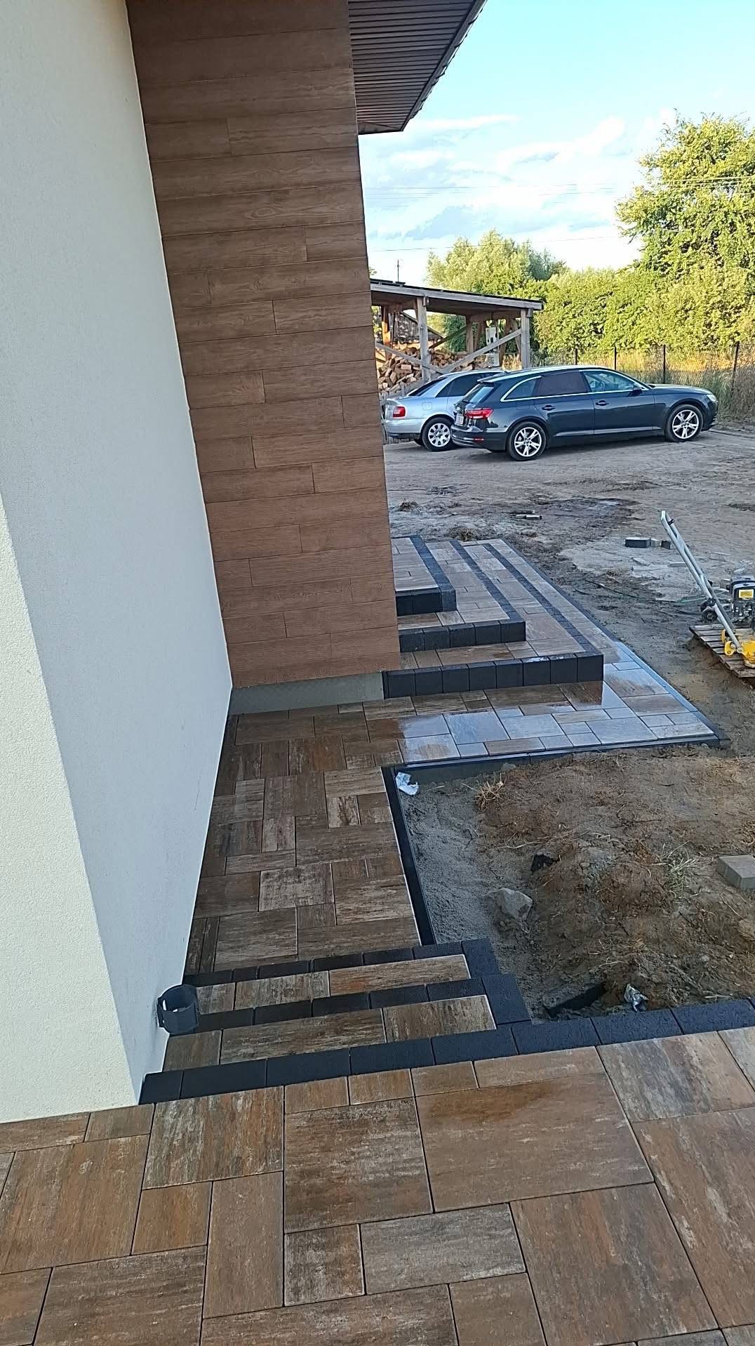 Ułożona kostka brukowa w odcieniach brązu i szarości tworząca schody i chodnik przed budynkiem. Widoczne dwa samochody w tle i częściowo niezakończone prace.