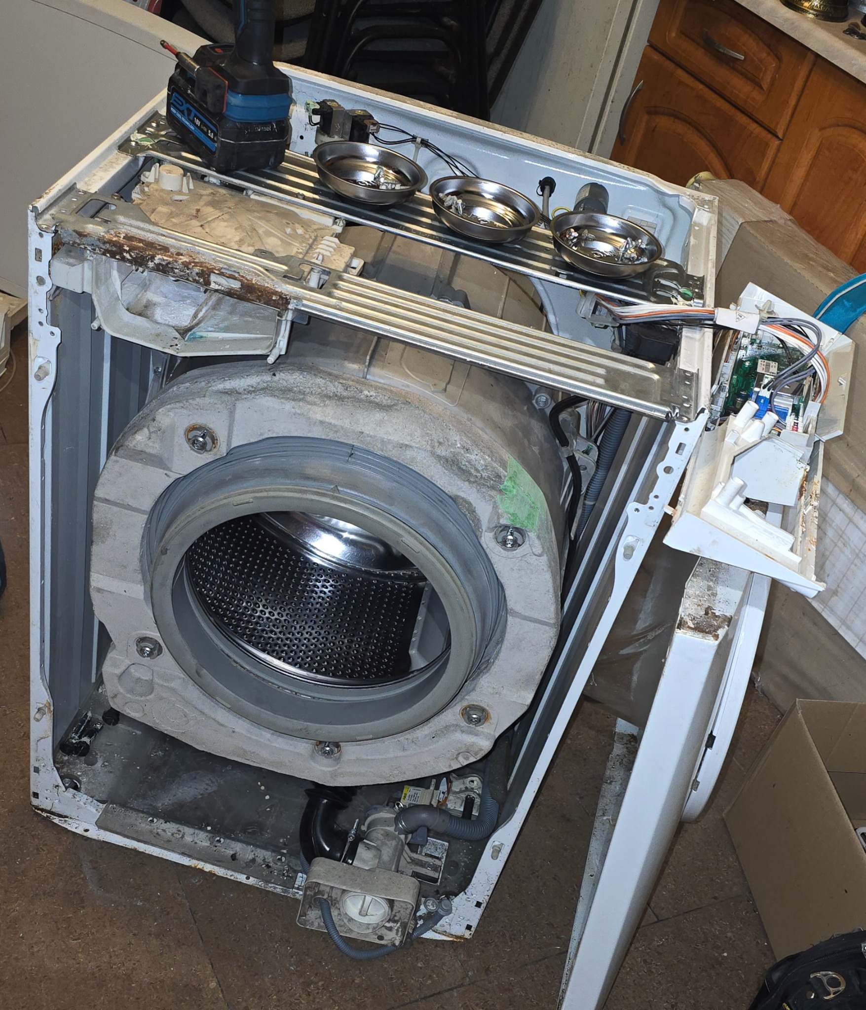 Rozłożona pralka automatyczna z widocznym bębnem, elementami elektronicznymi i narzędziami, sugerująca etap naprawy lub demontażu. Widoczne ślady użytkowania i zabrudzenia.