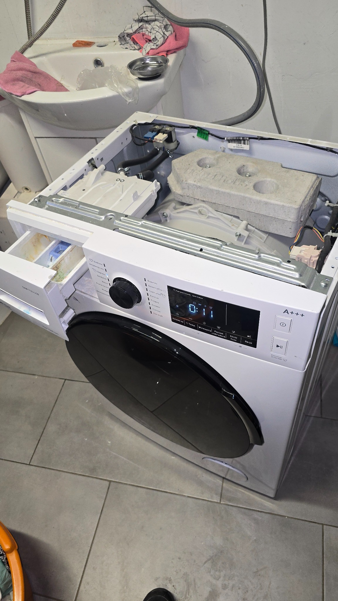 Otwarta pralka automatyczna z wysuniętą szufladą na proszek, widoczne elementy wewnętrzne i panel sterowania. W tle umywalka z akcesoriami.