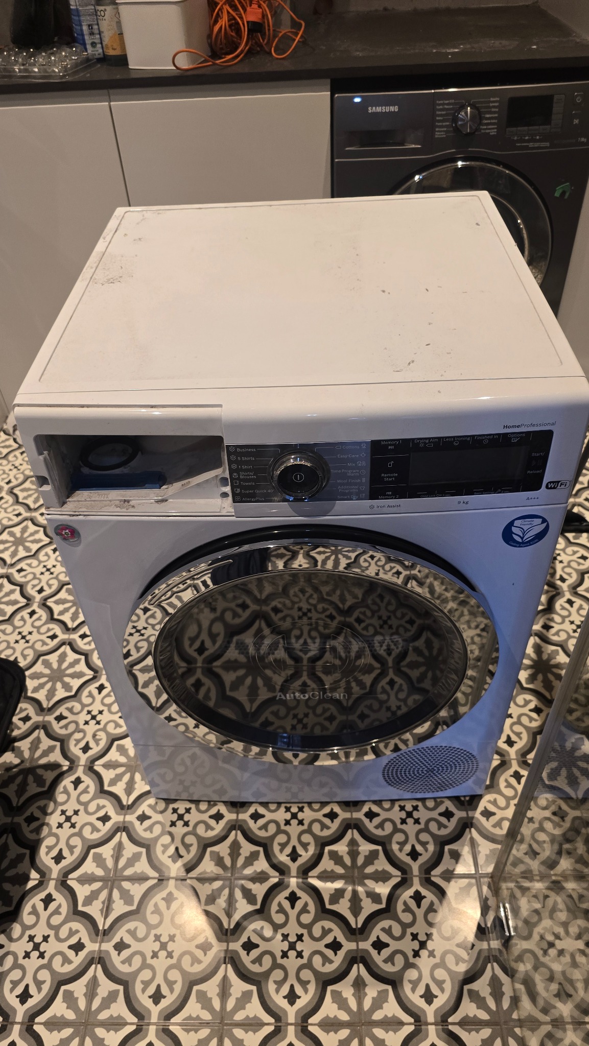 Biała suszarka HomeProfessional z otwartymi drzwiami na tle wzorzystej podłogi i pralki Samsung. Widoczny panel sterowania i oznaczenia energetyczne.