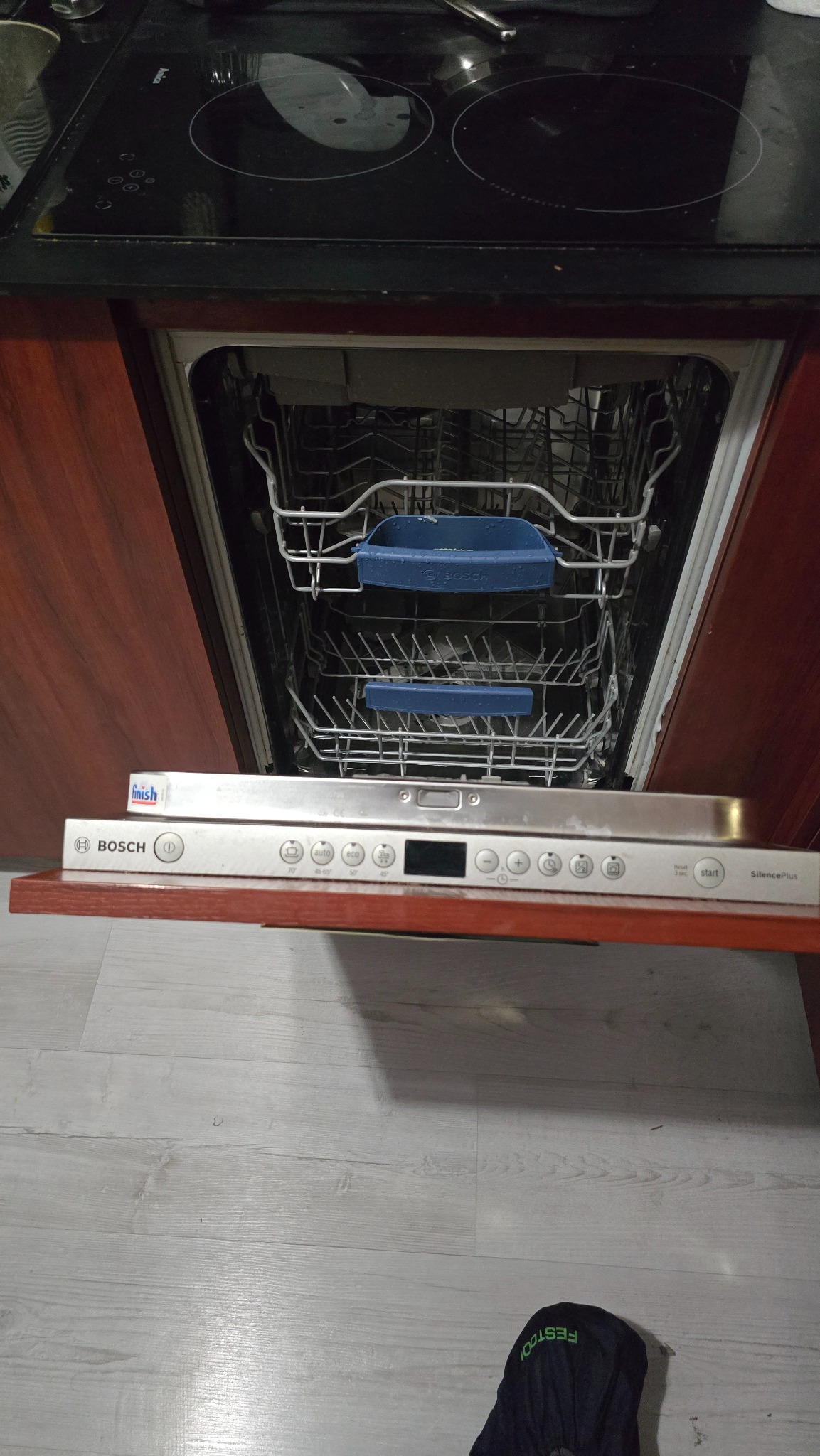 Otwarta zmywarka Bosch SilencePlus z widocznymi koszami na naczynia i panelem sterowania, umieszczona pod płytą indukcyjną w zabudowie kuchennej z drewnianymi frontami.