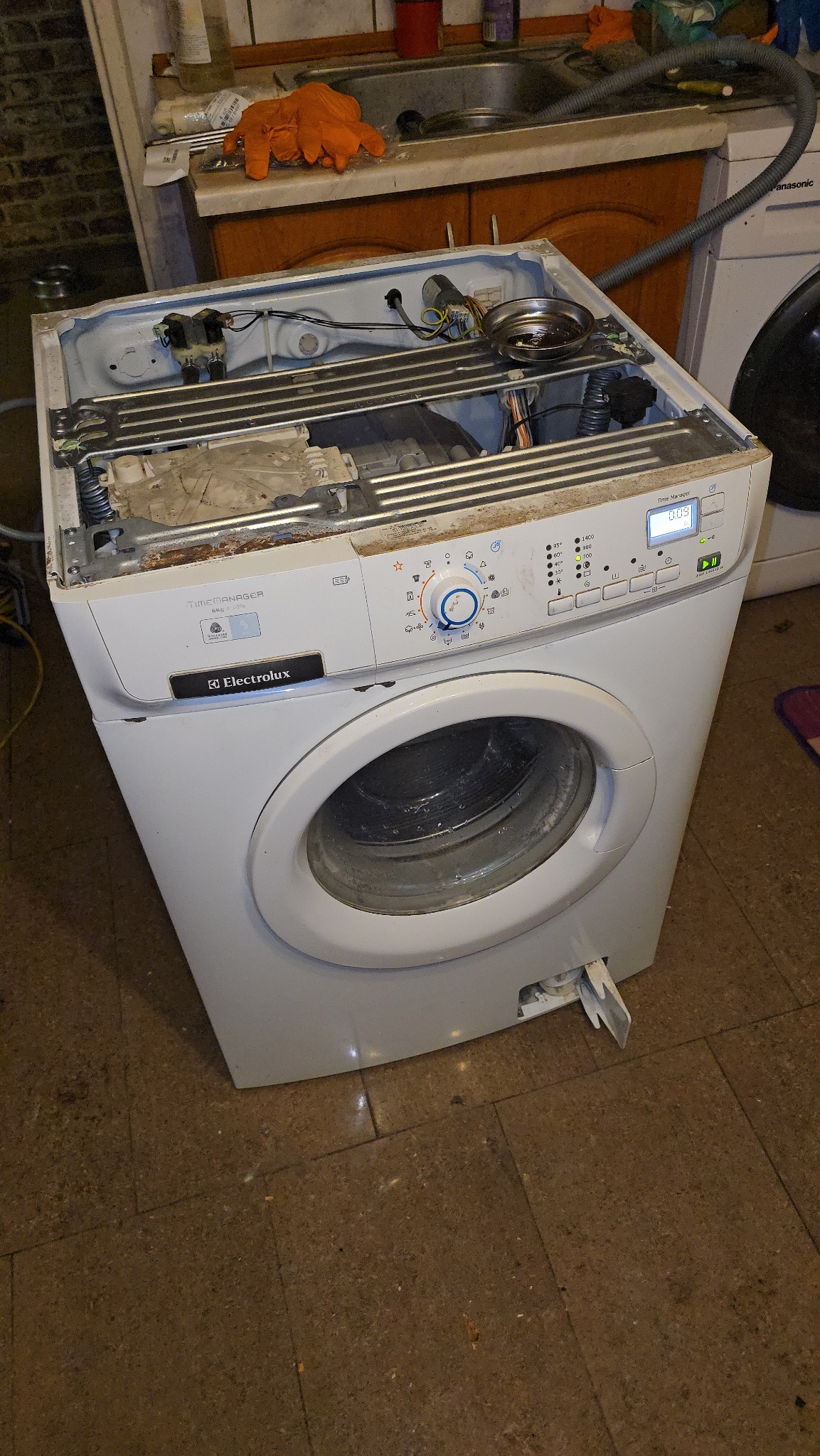 Rozłożona pralka Electrolux Time Manager 6kg z otwartą obudową, widoczne wnętrze i panel sterowania. W tle zlew i inne sprzęty AGD.