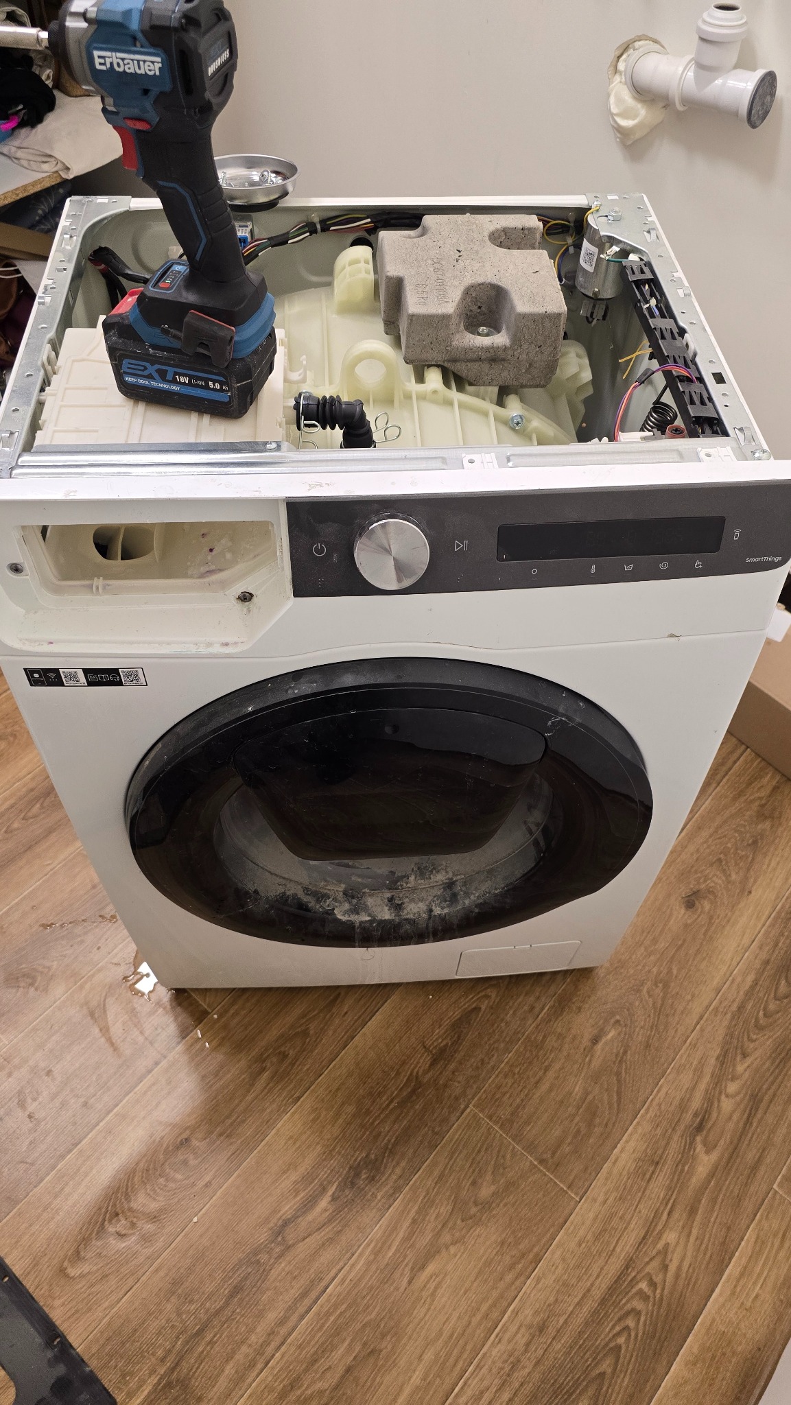 Otwarta pralka w trakcie naprawy, z widocznym wnętrzem i wkrętarką Erbauer na obudowie. Widoczne ślady użytkowania na obudowie i podłodze.