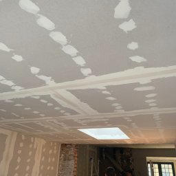 Tymchyna Expert Bud - Sufit z płyt kartonowo-gipsowych, z widocznymi łączeniami i szpachlowanymi miejscami po wkrętach, przygotowany do malowania. W tle fragment ściany z odsłoniętą cegłą.