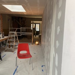Tymchyna Expert Bud - Wnętrze w trakcie prac wykończeniowych: ściany z płyt karton-gips, szpachlowane, z widocznymi łatami. Na podłodze folia ochronna, w tle rusztowanie i osoba.