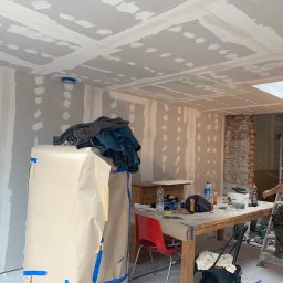 Tymchyna Expert Bud - Wnętrze w trakcie prac wykończeniowych w Markach: ściany i sufit z płyt karton-gips, szpachlowane łączenia, przygotowanie do malowania. Widoczny pracownik na drabinie.