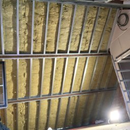 Tymchyna Expert Bud - Izolacja poddasza wełną mineralną, widoczne metalowe profile konstrukcyjne i fragment okna. Prace wykończeniowe w trakcie realizacji.