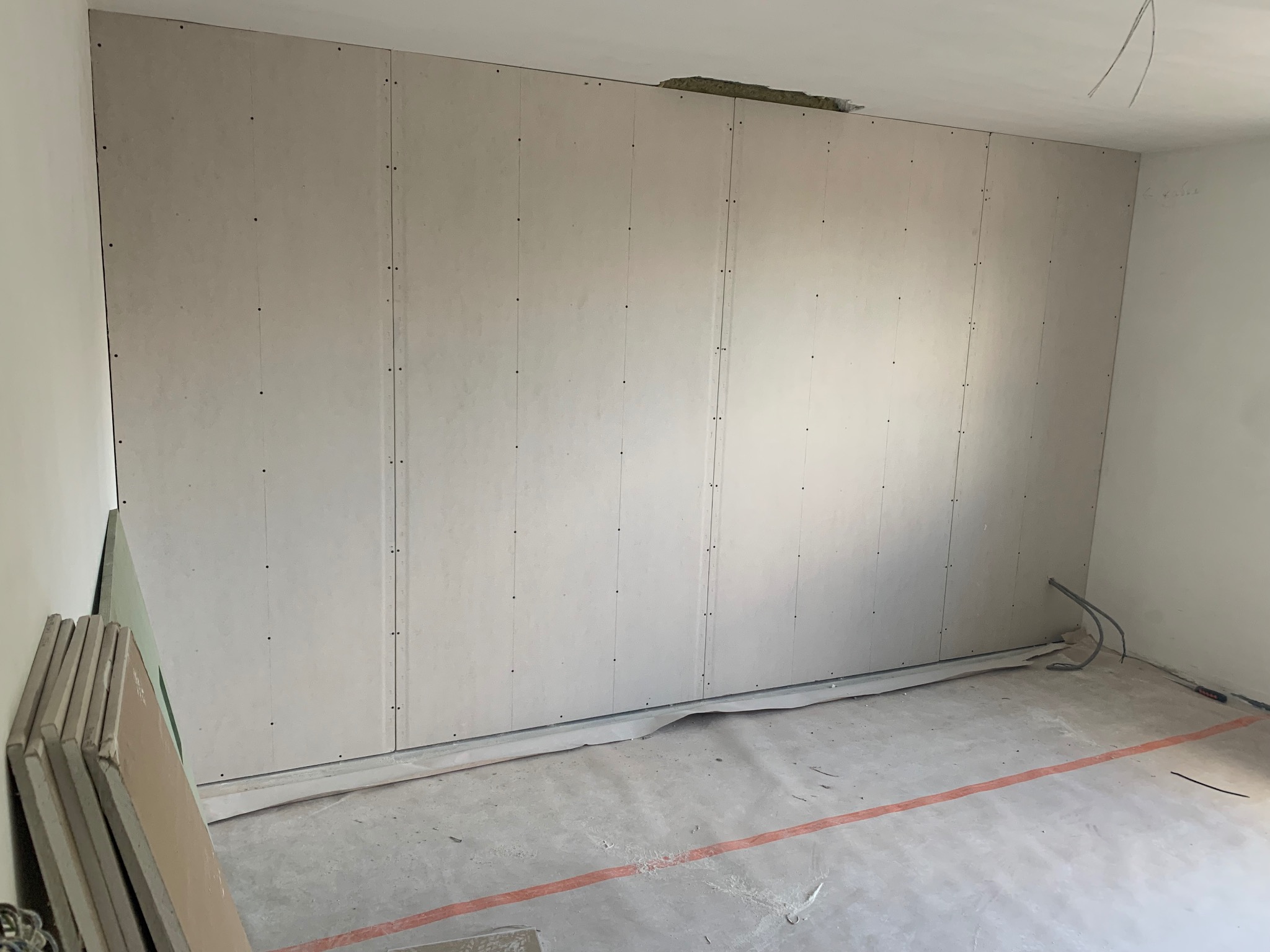 Ściana wykończona płytami kartonowo-gipsowymi, widoczne łączenia i wkręty. Na podłodze resztki materiałów budowlanych i przewody. Częściowo widoczne płyty gipsowe.