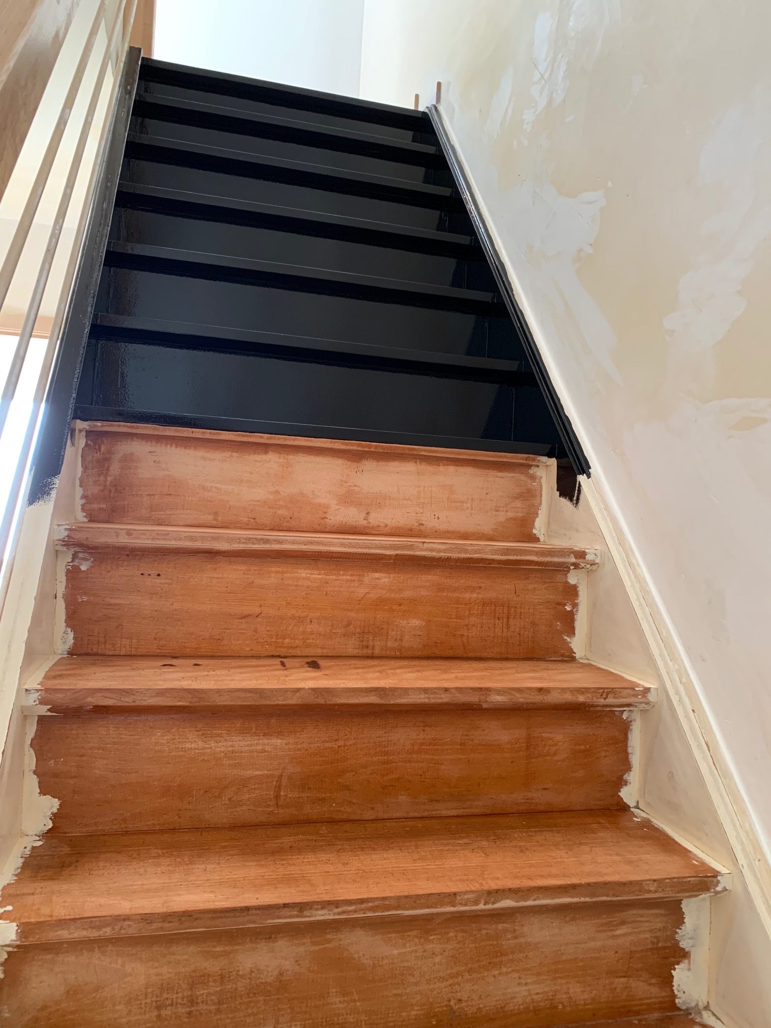 Renowacja schodów: dolne stopnie drewniane, przygotowane do malowania, górne pomalowane na czarno. Widoczne ślady szpachli na ścianie i niedomalowane fragmenty.