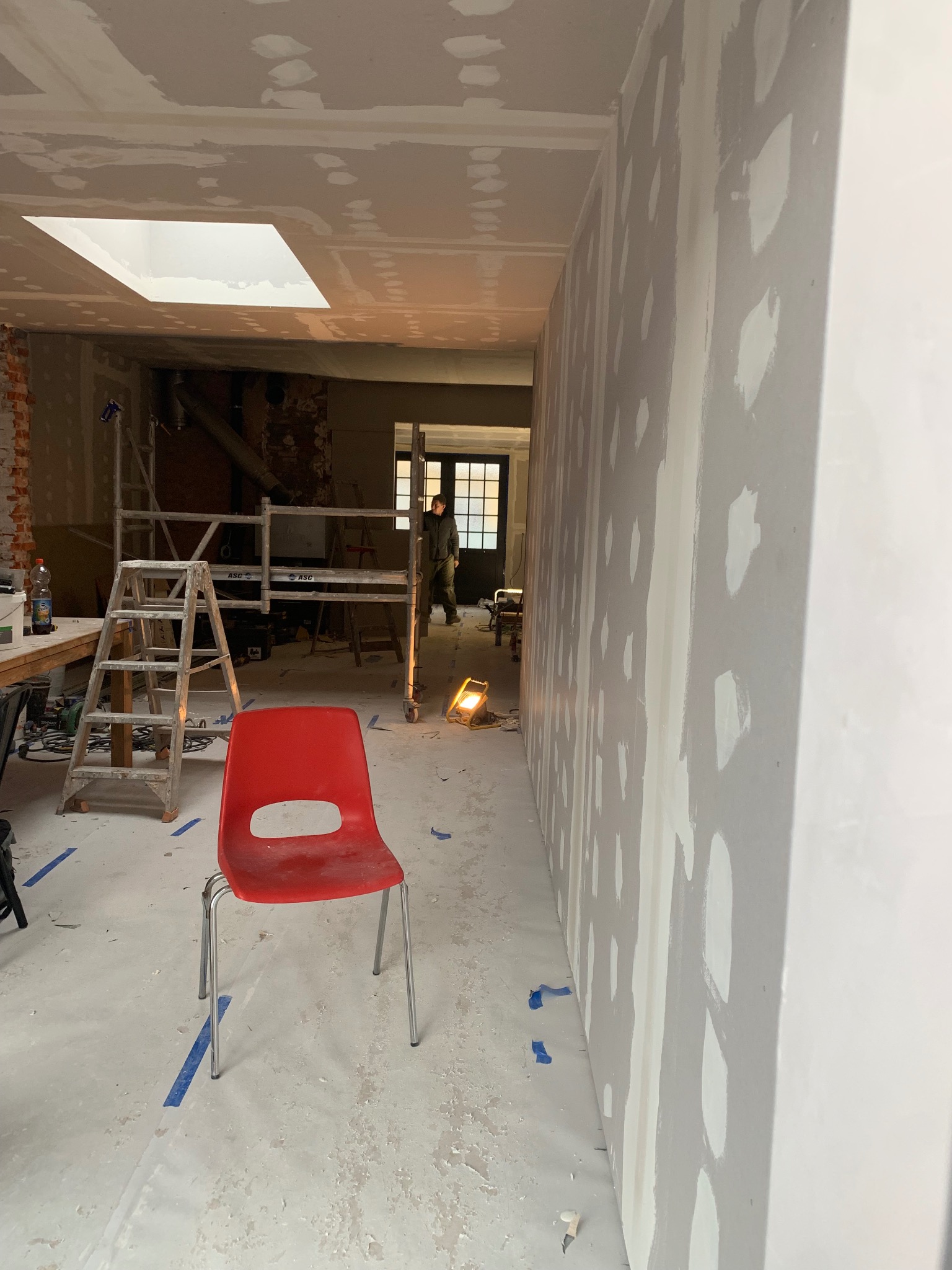 Wnętrze w trakcie prac wykończeniowych: ściany z płyt karton-gips, szpachlowane, z widocznymi łatami. Na podłodze folia ochronna, w tle rusztowanie i osoba.