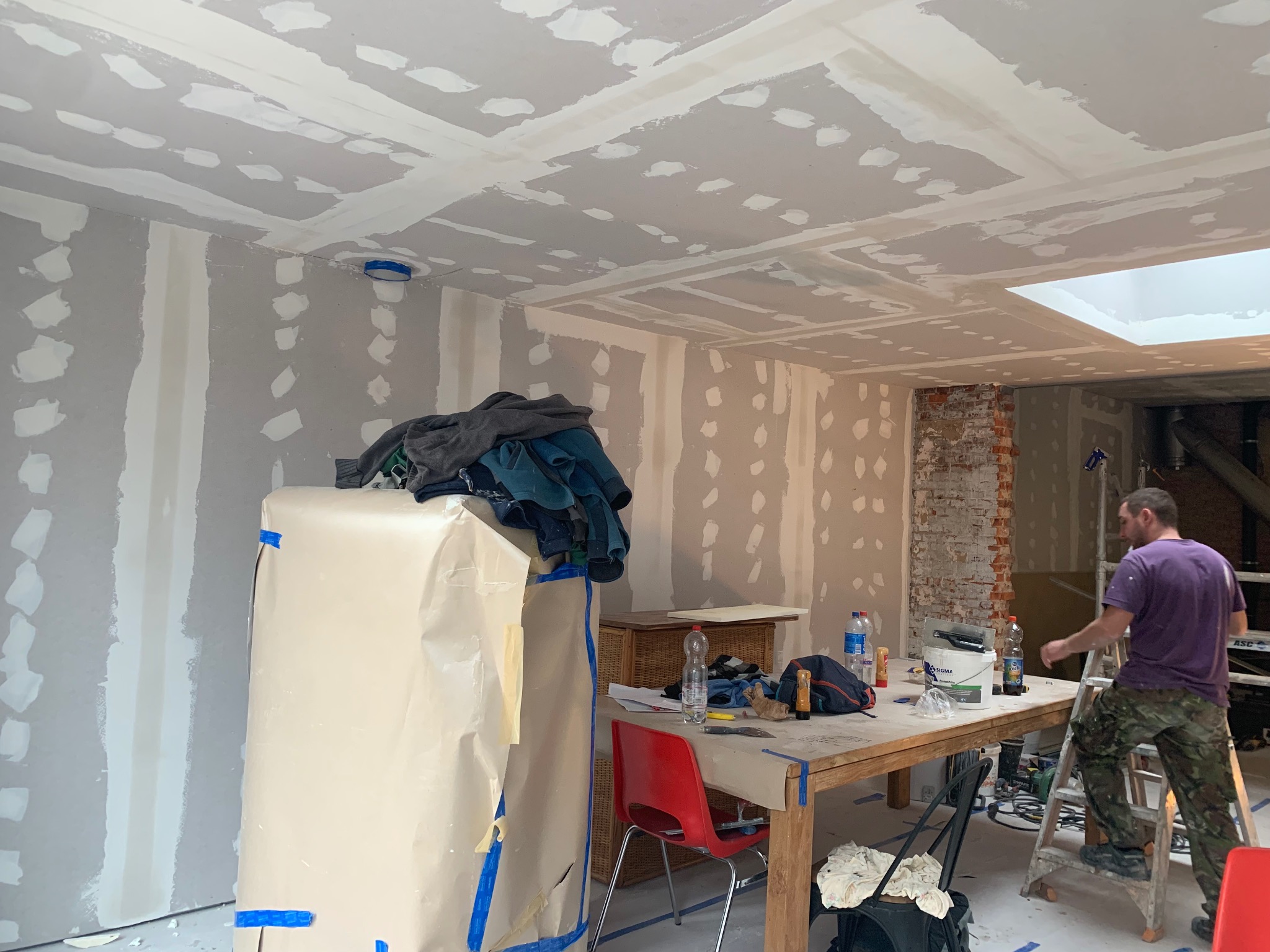 Wnętrze w trakcie prac wykończeniowych w Markach: ściany i sufit z płyt karton-gips, szpachlowane łączenia, przygotowanie do malowania. Widoczny pracownik na drabinie.