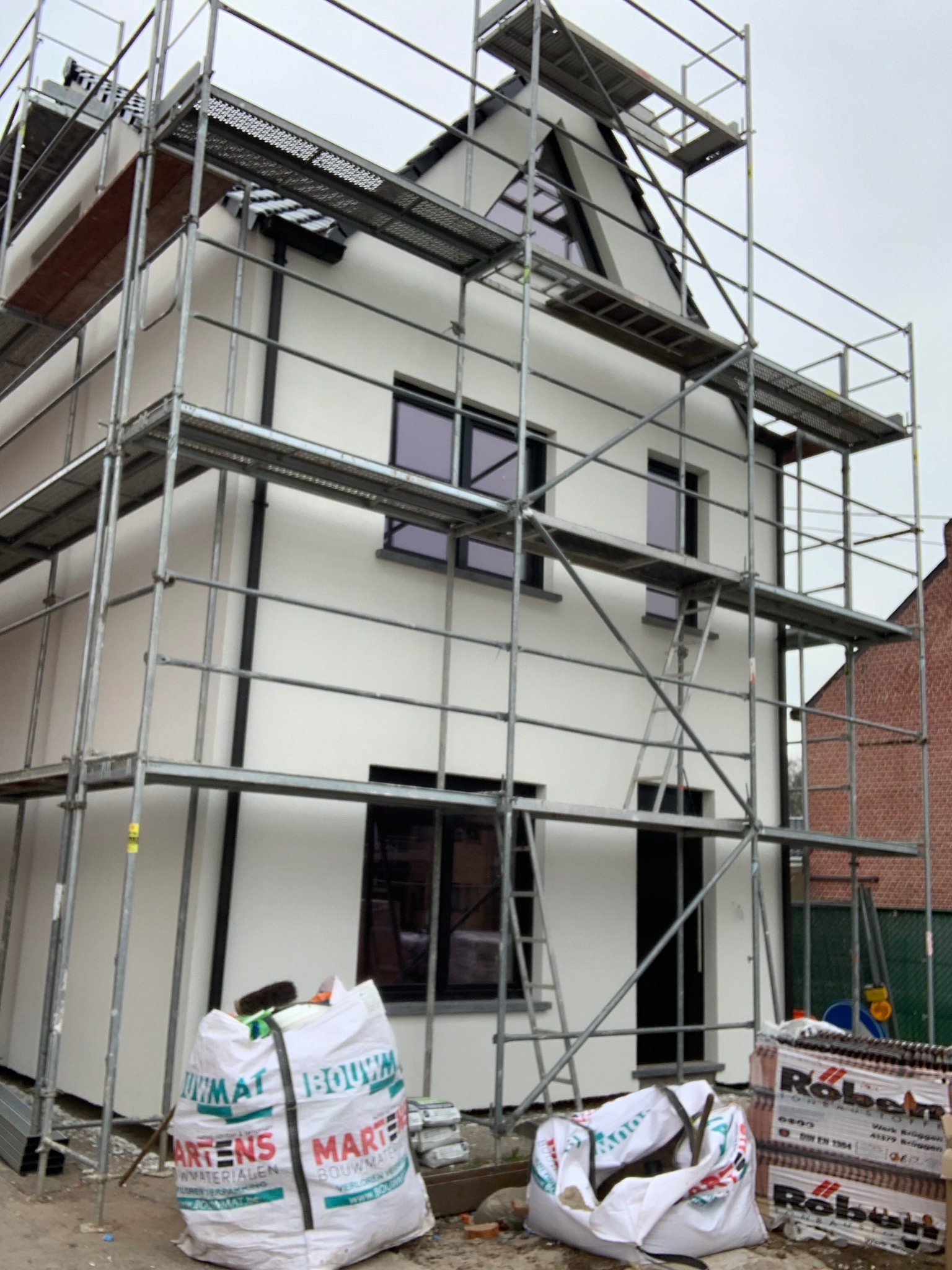 Nowoczesny dom w trakcie wykończenia elewacji, otoczony rusztowaniami. Widoczne worki z materiałami budowlanymi i cegły. Dom z białymi ścianami i ciemnymi oknami.