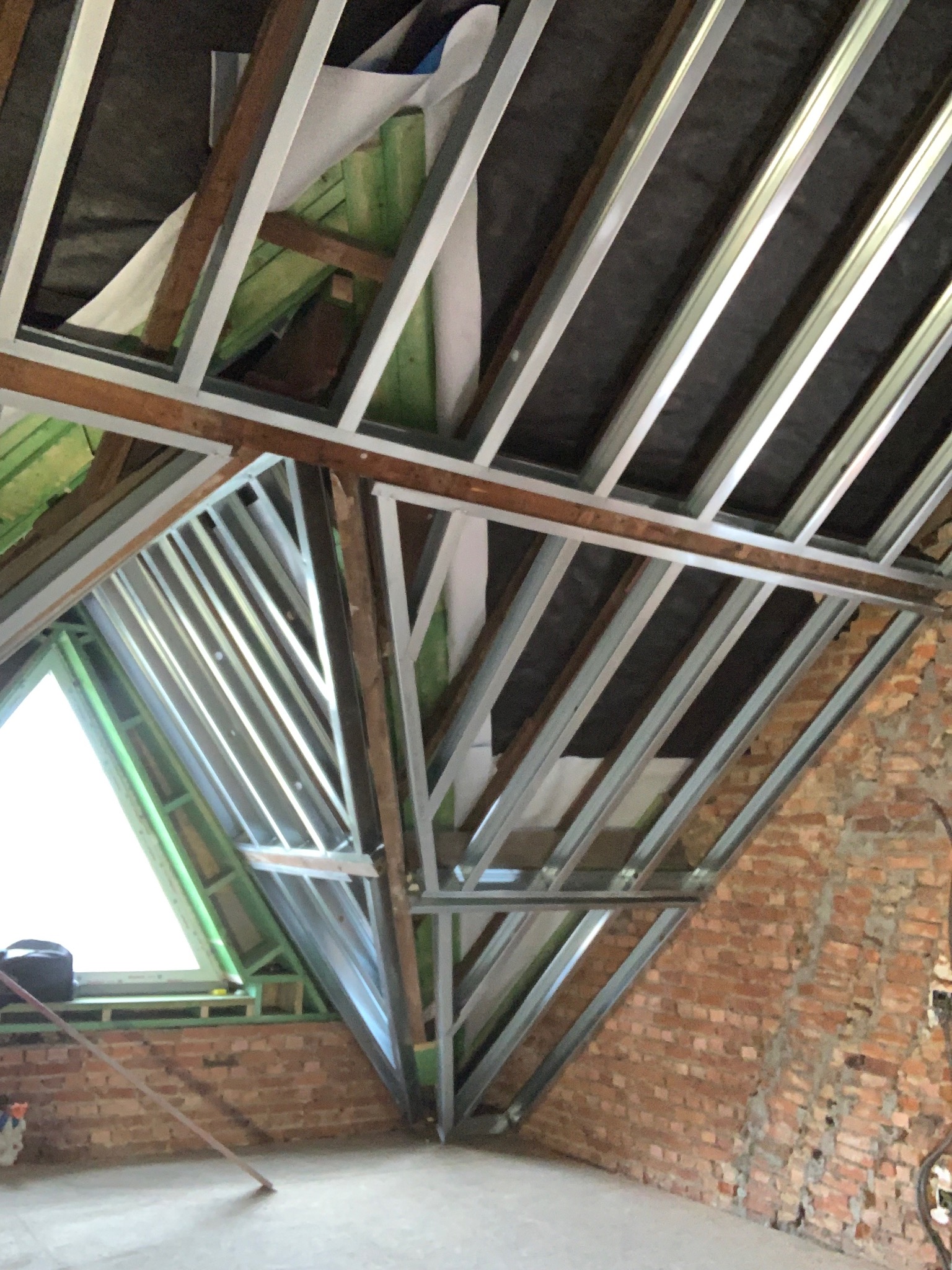 Szkielet metalowy poddasza z oknem, widoczne drewniane elementy konstrukcyjne i fragment ceglanej ściany. Prace wykończeniowe w trakcie realizacji.