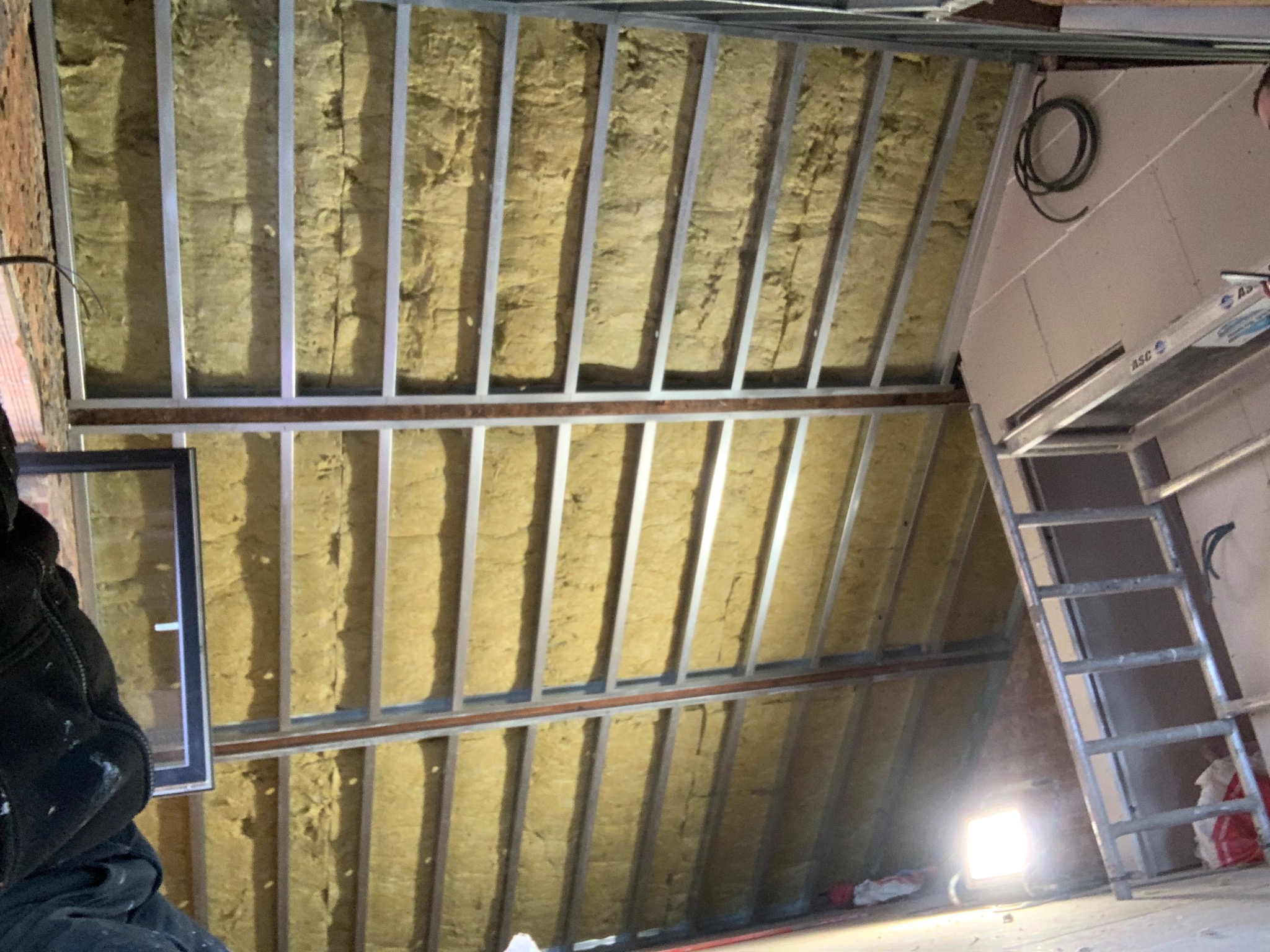 Izolacja poddasza wełną mineralną, widoczne metalowe profile konstrukcyjne i fragment okna. Prace wykończeniowe w trakcie realizacji.