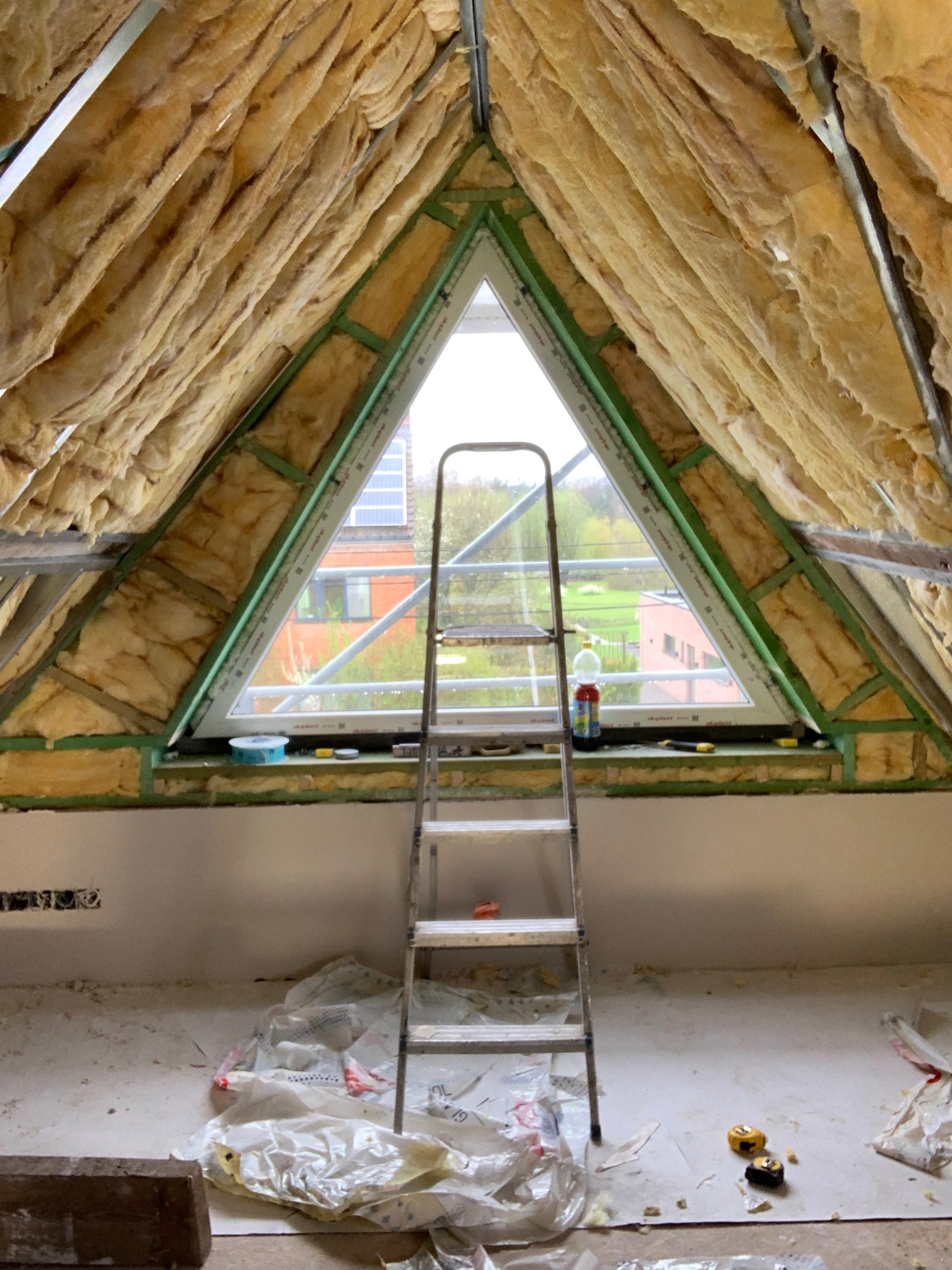 Poddasze z oknem w kształcie trójkąta, ocieplone wełną mineralną, z drabinką opartą o parapet. Widoczne narzędzia i materiały budowlane. Etap prac wykończeniowych.