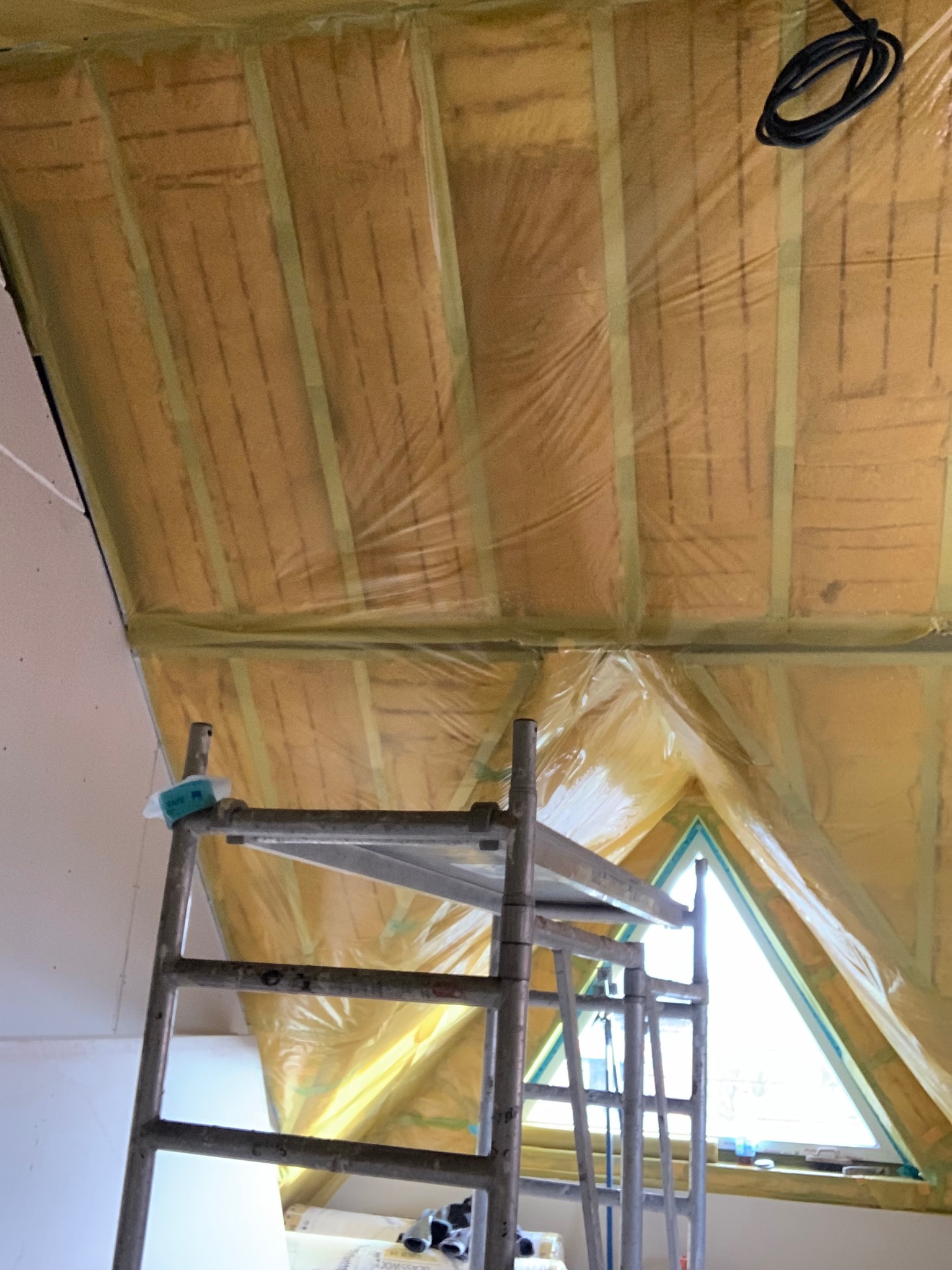 Ocieplenie poddasza wełną mineralną, zabezpieczone folią, widoczne z rusztowania. Okno trójkątne w tle. Prace wykończeniowe w trakcie realizacji.