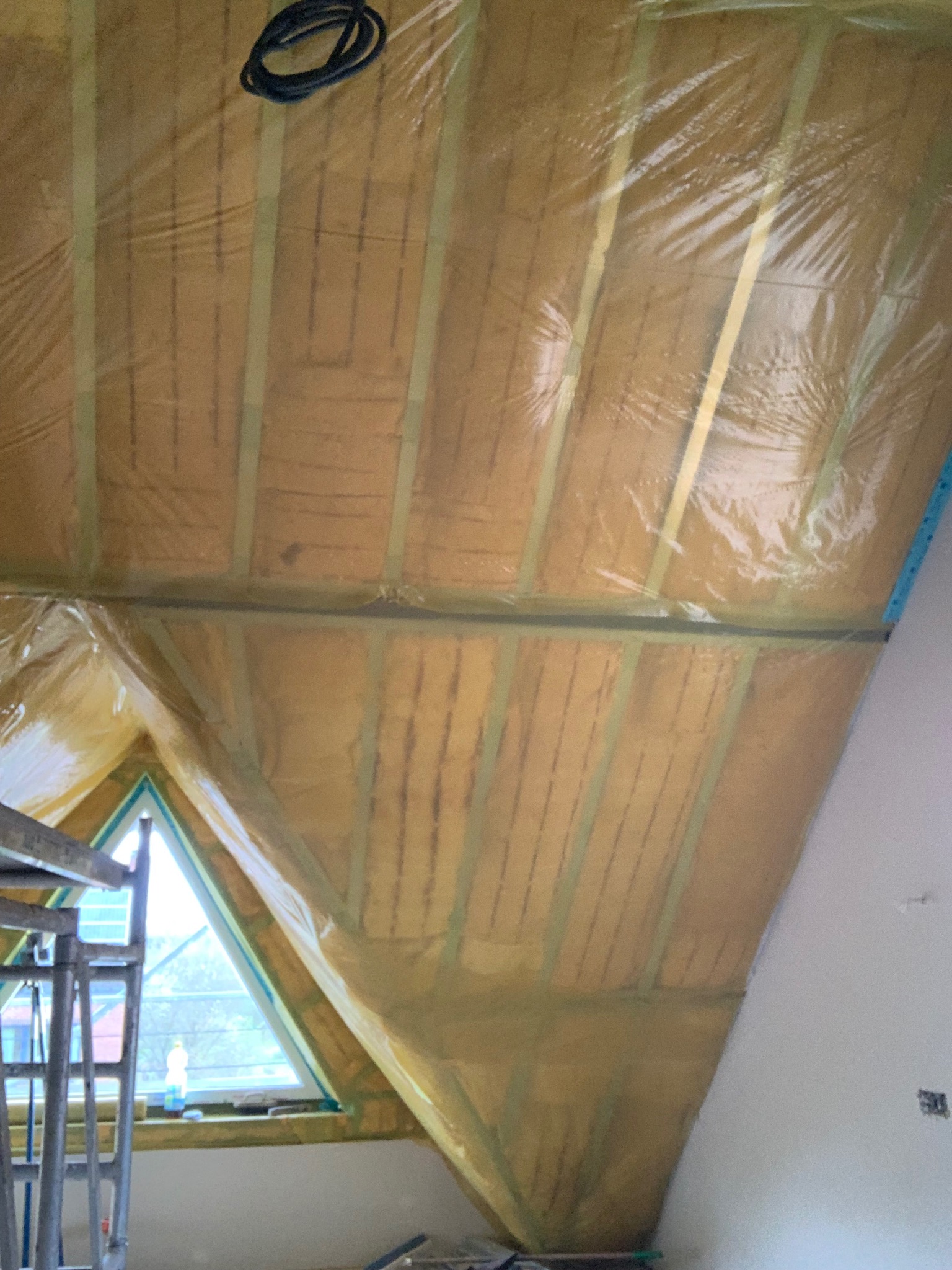 Ocieplenie poddasza wełną mineralną, zabezpieczone folią paroizolacyjną. Widoczne drewniane krokwie i fragment okna dachowego. Prace wykończeniowe w trakcie.