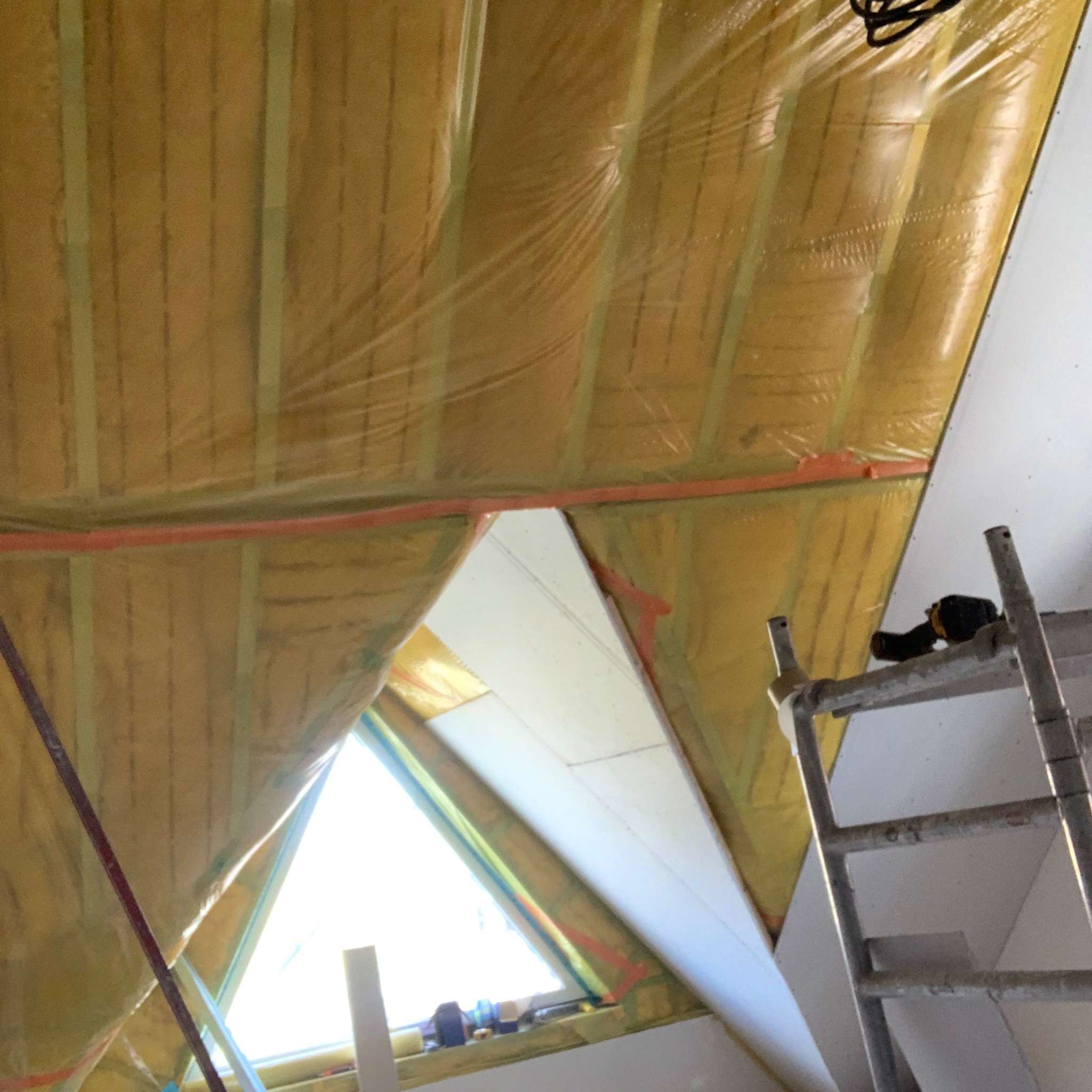 Ocieplenie poddasza wełną mineralną zabezpieczoną folią, widoczne płyty karton-gips i drabina z wkrętarką. Prace wykończeniowe w trakcie adaptacji poddasza.