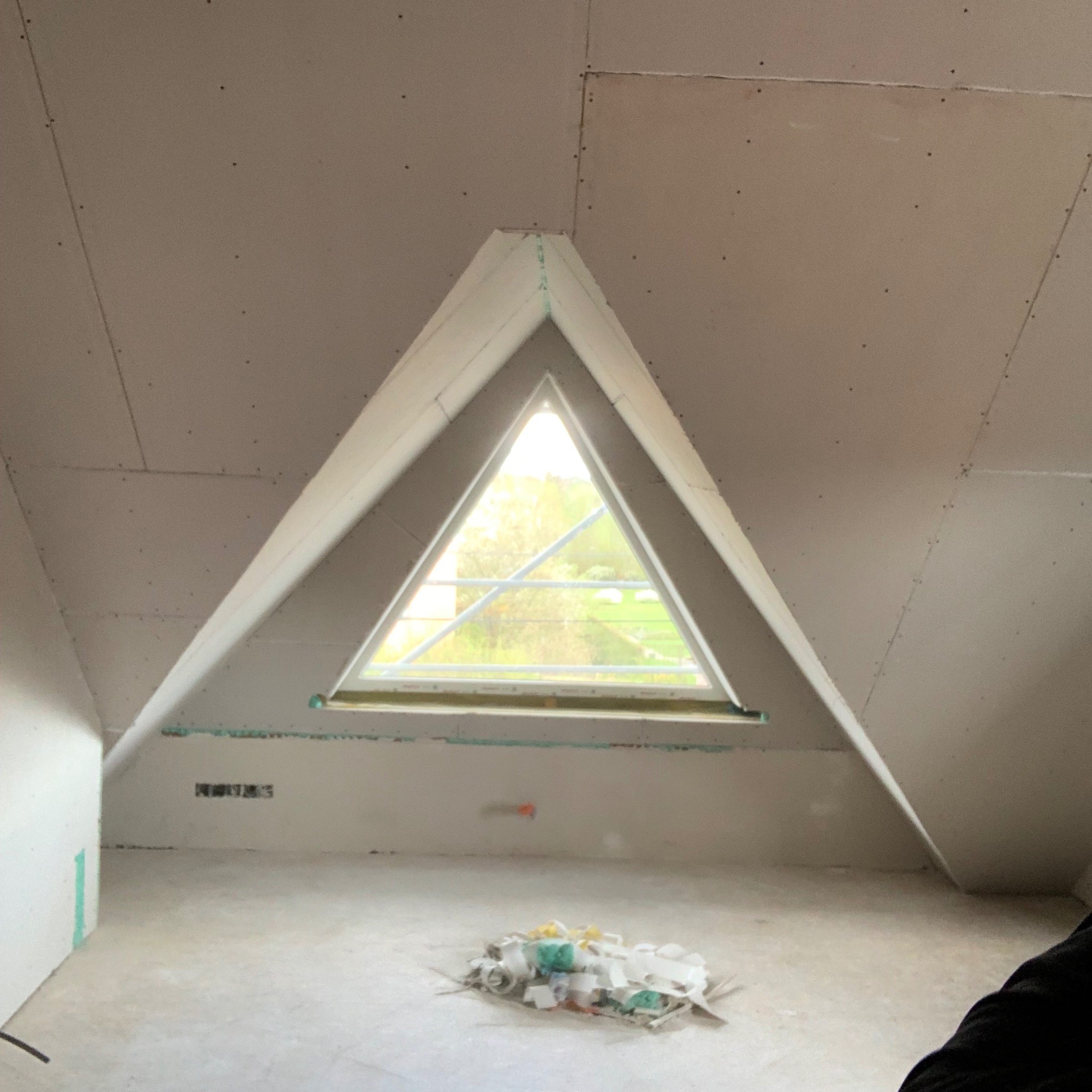 Poddasze z oknem w kształcie trójkąta, ściany i sufit wykończone płytami kartonowo-gipsowymi, widoczne łączenia. Na podłodze resztki materiałów budowlanych.