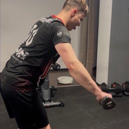 Treningi personalne - Jędrzej Niepokój - Mężczyzna w sportowym stroju wykonuje ćwiczenie z hantlą w domowej siłowni, widoczny sprzęt do ćwiczeń i robot sprzątający.