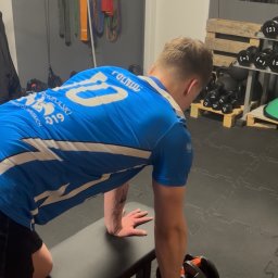 Treningi personalne - Jędrzej Niepokój - Mężczyzna w niebieskiej koszulce ćwiczy z hantlami na ławeczce w siłowni. Widoczne gumy do ćwiczeń i regał z obciążeniami w tle.