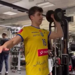 Treningi personalne - Jędrzej Niepokój - Mężczyzna w sportowej koszulce z numerem 16 ćwiczy z hantlą na siłowni w Bydgoszczy. W tle widoczne maszyny do ćwiczeń i inne osoby.