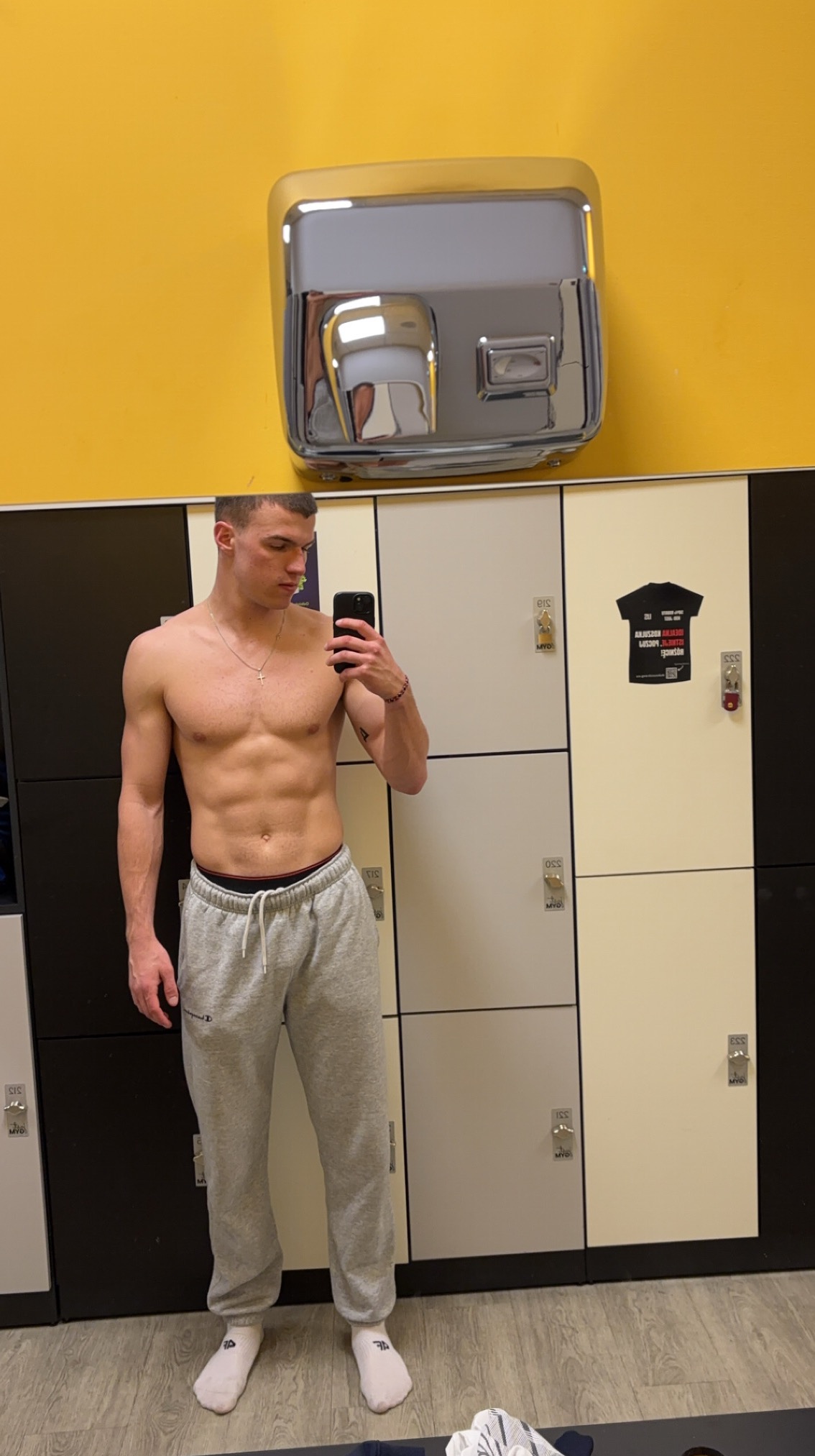 Mężczyzna bez koszulki robi selfie w szatni siłowni, prezentując wyrzeźbione ciało na tle szafek i suszarki do rąk. Widoczny tatuaż na ramieniu i łańcuszek z krzyżykiem.