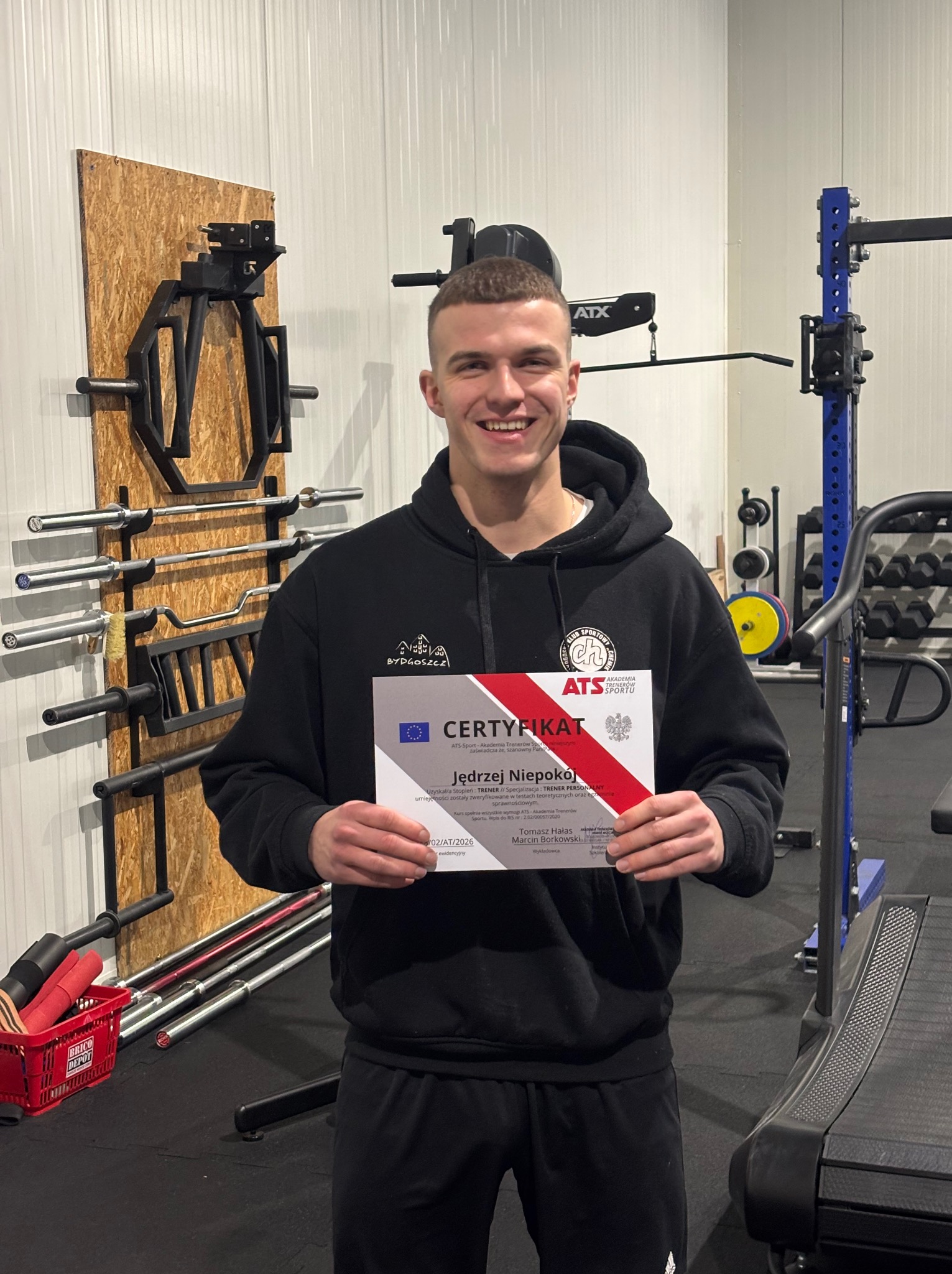 Uśmiechnięty mężczyzna w bluzie prezentuje certyfikat trenera personalnego na tle siłowni z bieżnią i sprzętem do ćwiczeń siłowych.