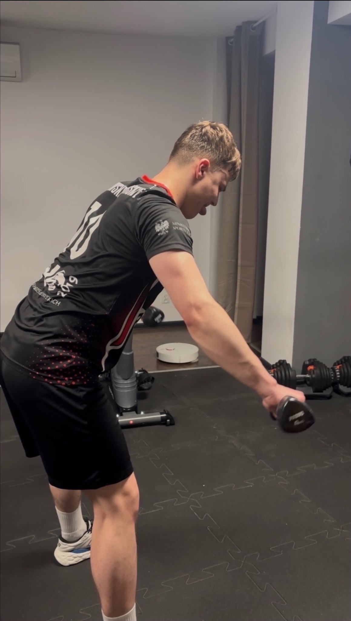 Mężczyzna w sportowym stroju wykonuje ćwiczenie z hantlą w domowej siłowni, widoczny sprzęt do ćwiczeń i robot sprzątający.