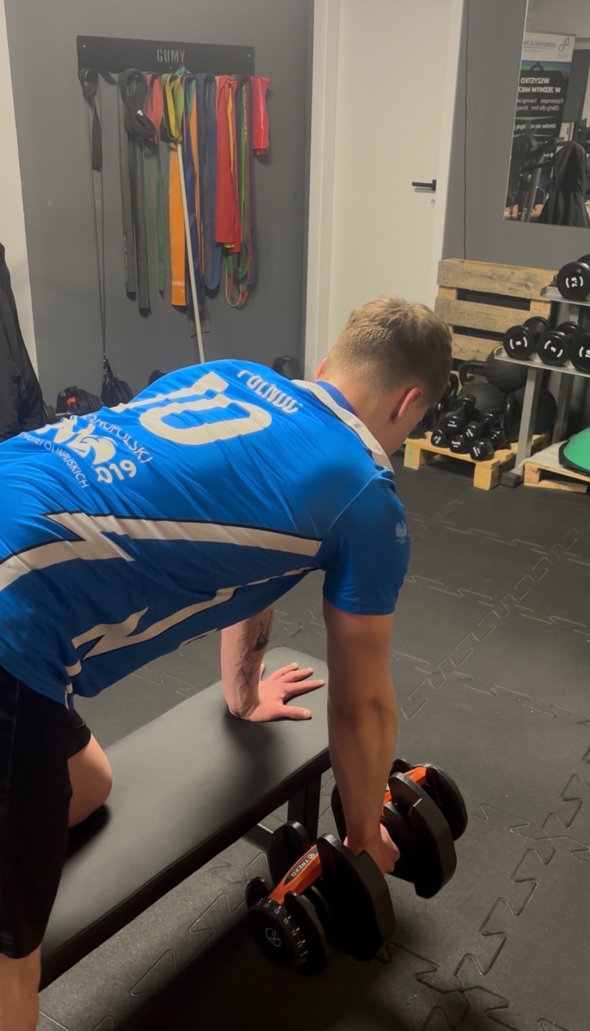 Mężczyzna w niebieskiej koszulce ćwiczy z hantlami na ławeczce w siłowni. Widoczne gumy do ćwiczeń i regał z obciążeniami w tle.