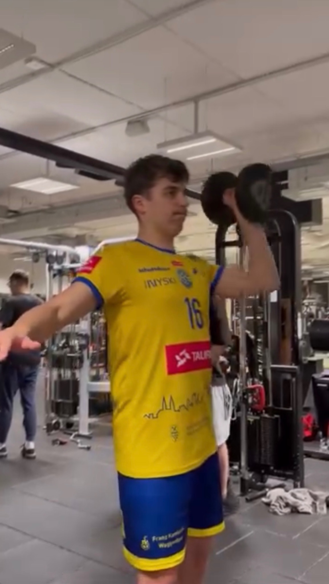 Mężczyzna w sportowej koszulce z numerem 16 ćwiczy z hantlą na siłowni w Bydgoszczy. W tle widoczne maszyny do ćwiczeń i inne osoby.