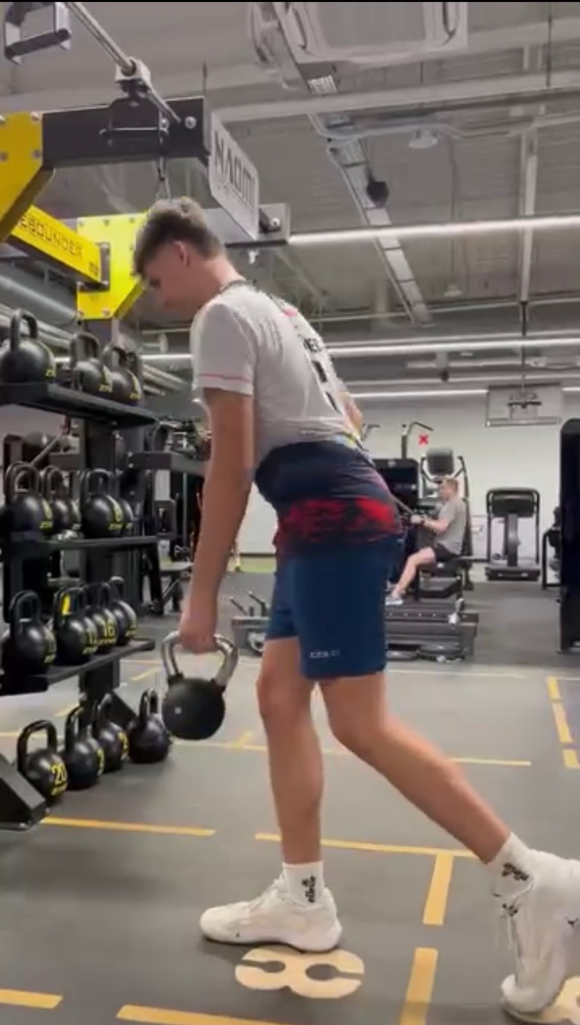 Młody mężczyzna w sportowym stroju wykonuje ćwiczenie z kettlebell w nowoczesnej siłowni. Widoczne regały z obciążeniami i sprzęt do ćwiczeń.