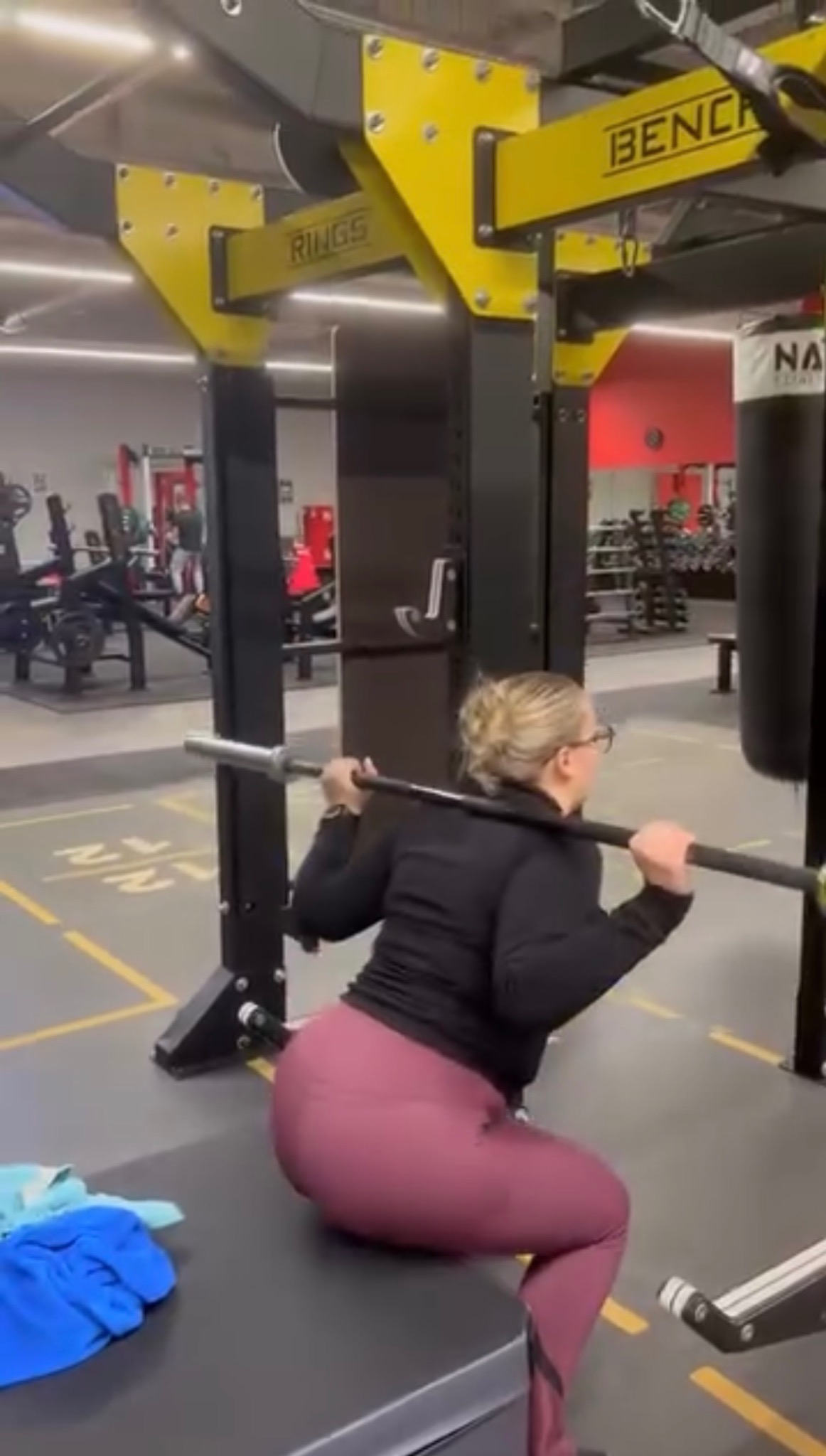 Kobieta w okularach wykonuje przysiad ze sztangą na ławeczce w siłowni. Żółto-czarna konstrukcja do ćwiczeń, worek bokserski i sprzęt w tle.
