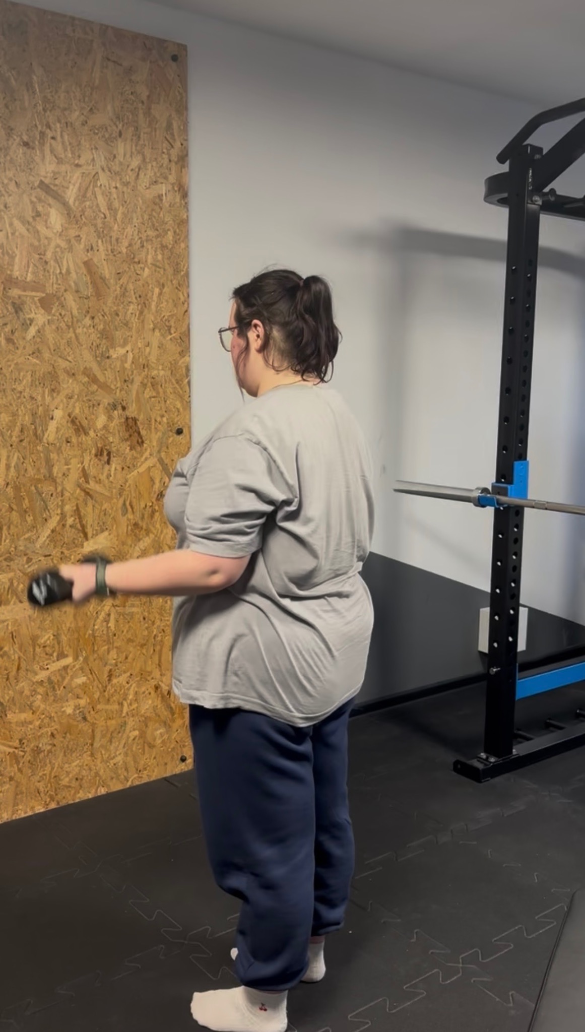 Kobieta w szarej koszulce ćwiczy z hantlami w siłowni. W tle ściana z płyty OSB i sprzęt do ćwiczeń siłowych. Widok z tyłu, skupienie na technice wykonywania ćwiczenia.
