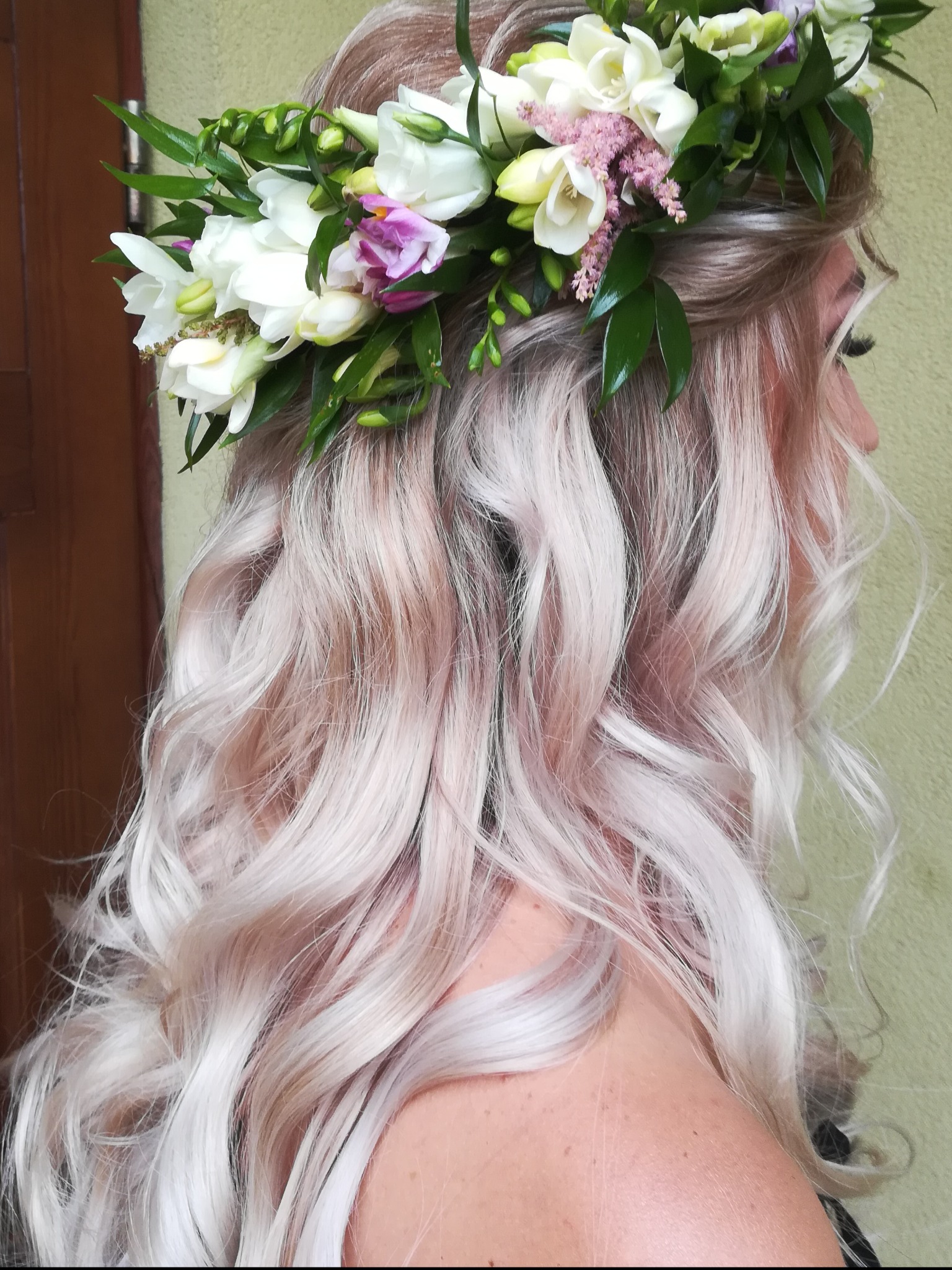 Profil kobiety z platynowo-różowymi, kręconymi włosami upiętymi w wianek z białych i fioletowych kwiatów oraz zielonych liści, na tle ściany i fragmentu brązowych drzwi.