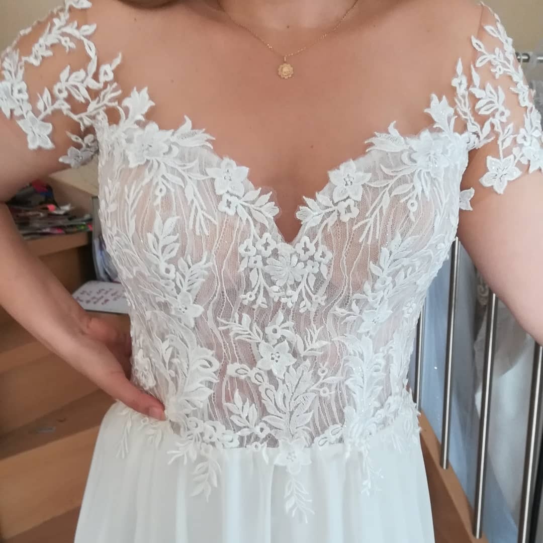 Biała suknia ślubna z koronkowym gorsetem i tiulowym dołem, widoczna na modelce, z delikatną złotą zawieszką na szyi, sfotografowana w naturalnym świetle.