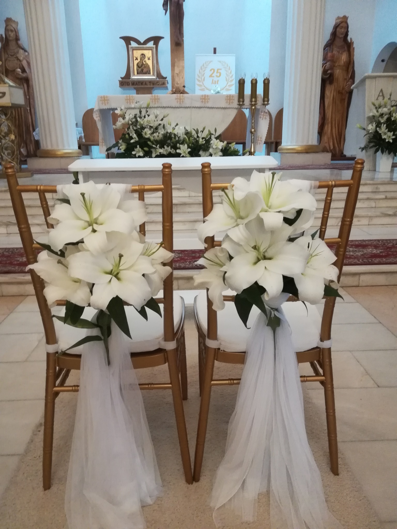 Uroczyste dekoracje z białych lilii i tiulu na złotych krzesłach w kościele, przygotowane na ceremonię ślubną.