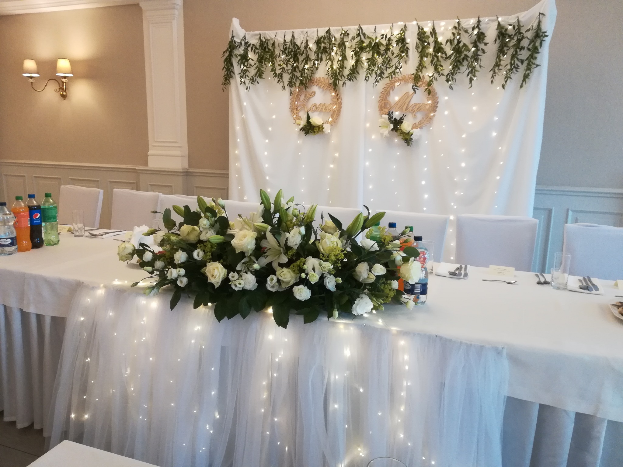 Uroczysty stół weselny z białym obrusem, dekoracją z kwiatów w odcieniach bieli i zieleni, podświetlonym tiulem, w tle biała ściana z girlandami i drewnianymi dekoracjami z imionami.