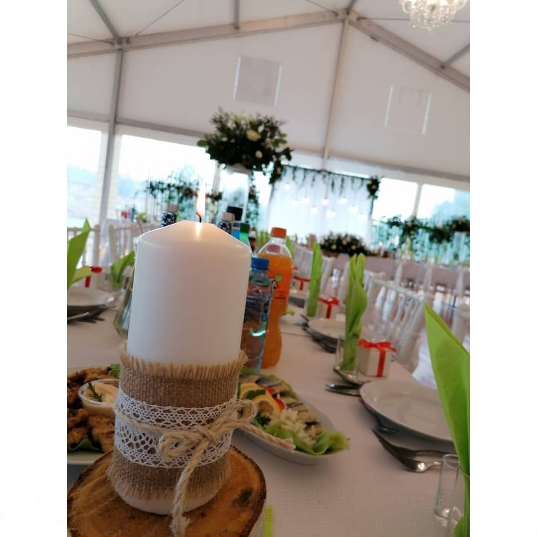 Dekoracja stołu weselnego: świeca w jutowym oplocie z koronką, ustawiona na drewnianym plastrze, z widocznymi potrawami i napojami, w tle białe krzesła i dekoracje kwiatowe.