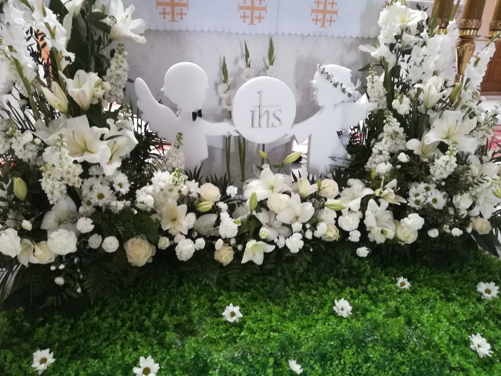 Dekoracja kwiatowa z białych lilii, róż i margerytek, z dekoracyjnymi figurkami aniołów trzymających okrągły znak IHS, na tle kościelnego ołtarza.