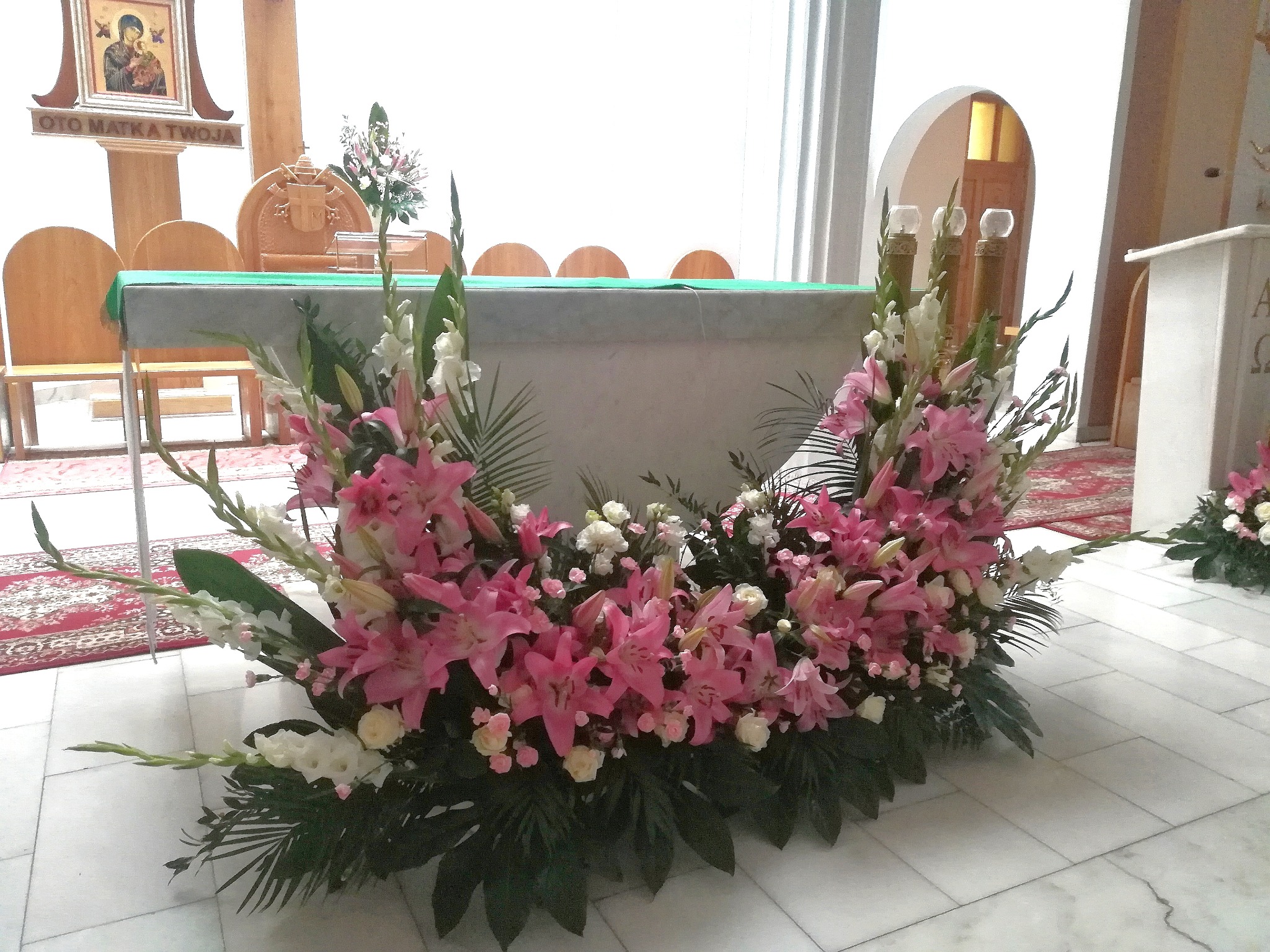 Dekoracja kwiatowa z różowych lilii i białych goździków na marmurowej posadzce przed ołtarzem w kościele, widoczne ikona religijna i krzesła z zielonym obrusem.