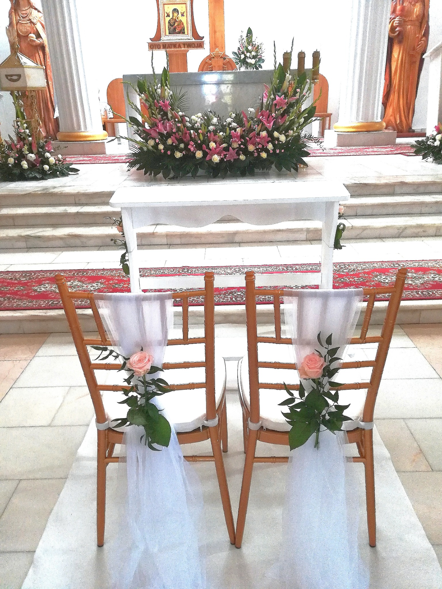 Uroczysta dekoracja kościoła na ślub: kwiaty na ołtarzu i krzesła udekorowane tiulem i różami w kolorze brzoskwini.