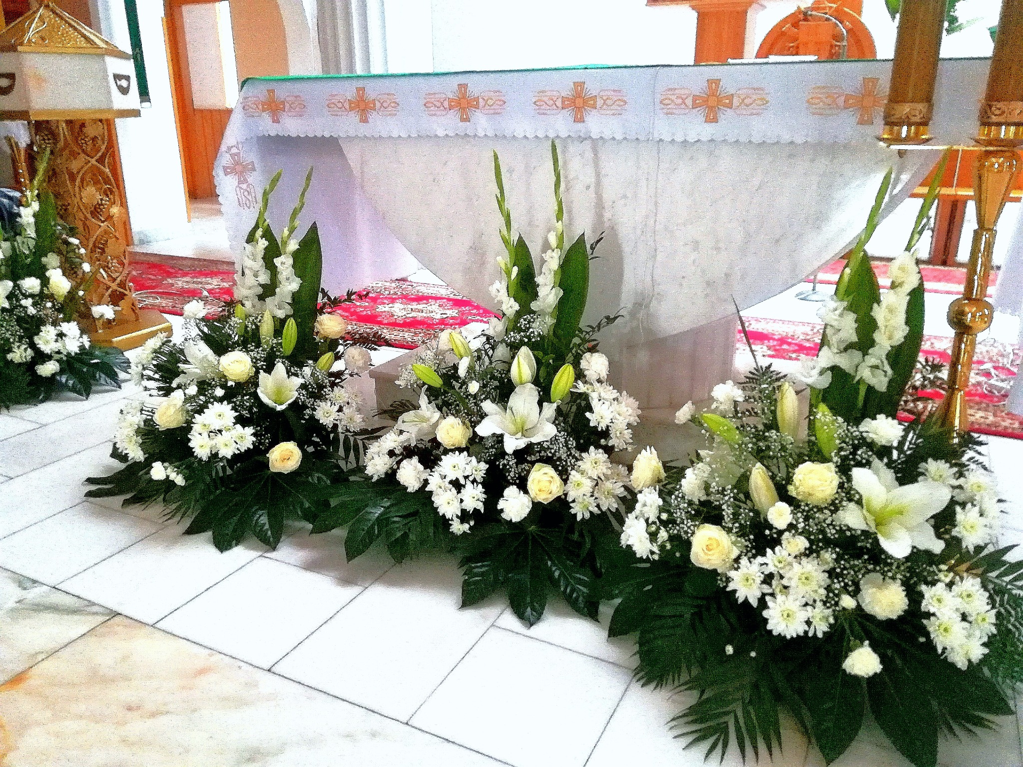 Dekoracja kwiatowa z białych róż i lilii, zdobiąca ołtarz w kościele, z widocznymi elementami wystroju wnętrza.