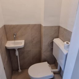 KAMIL SULOWSKI - Mała łazienka po remoncie: biała toaleta i umywalka narożna na tle beżowych płytek. Widoczny fragment drzwi. Minimalistyczny design, czystość i funkcjonalność.