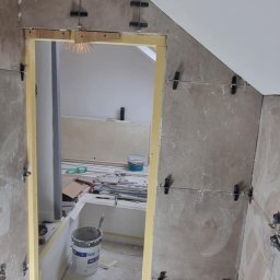 KAMIL SULOWSKI - Ściana w trakcie wykańczania płytkami, z użyciem klipsów poziomujących. Widoczne wiaderka i narzędzia w tle, sufit ze skosem.