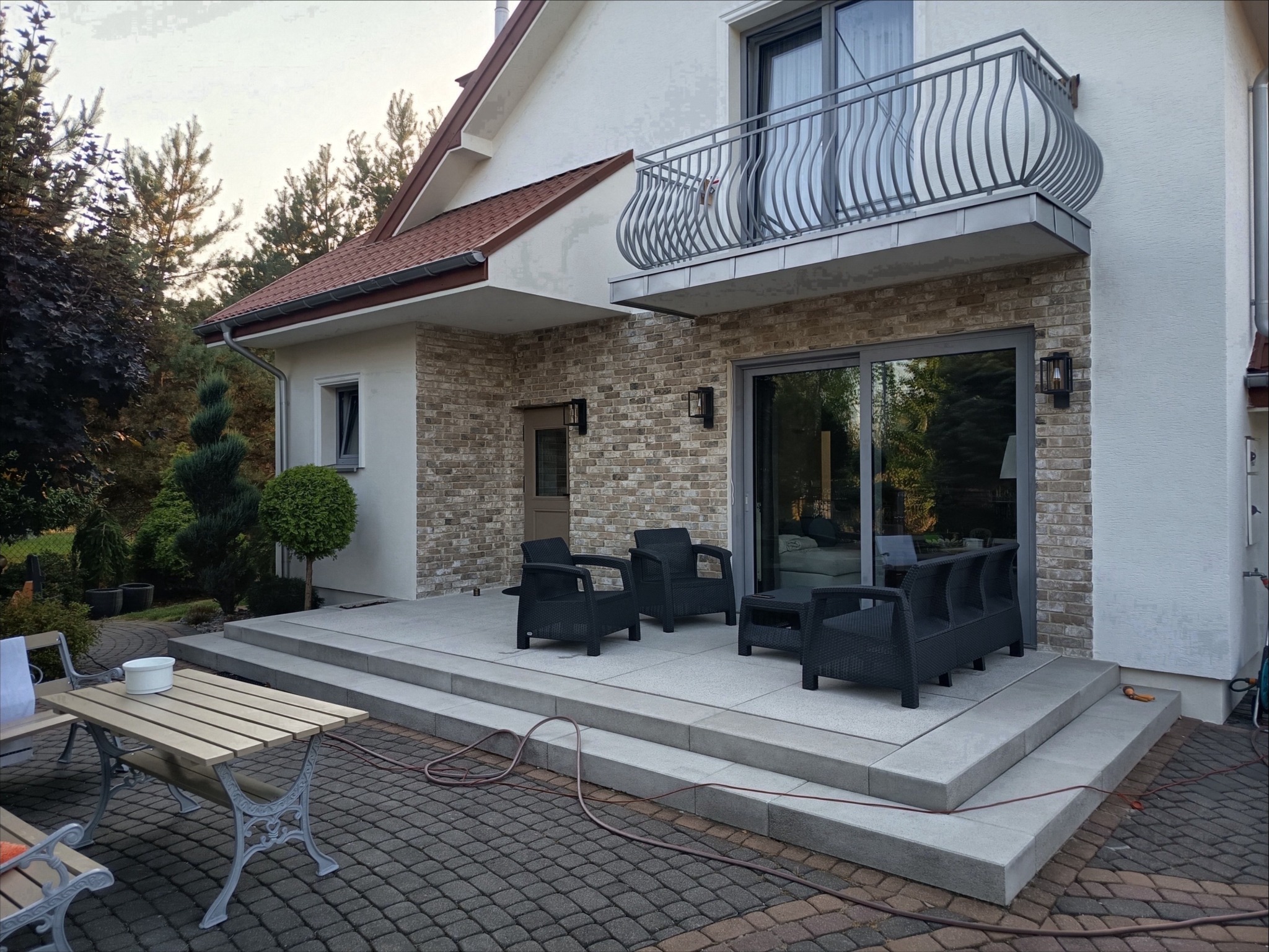 Elegancki taras z szarej kostki brukowej i meblami ogrodowymi, z widokiem na balkon z ozdobną balustradą. Dom z ceglaną elewacją i nowoczesnymi lampami.
