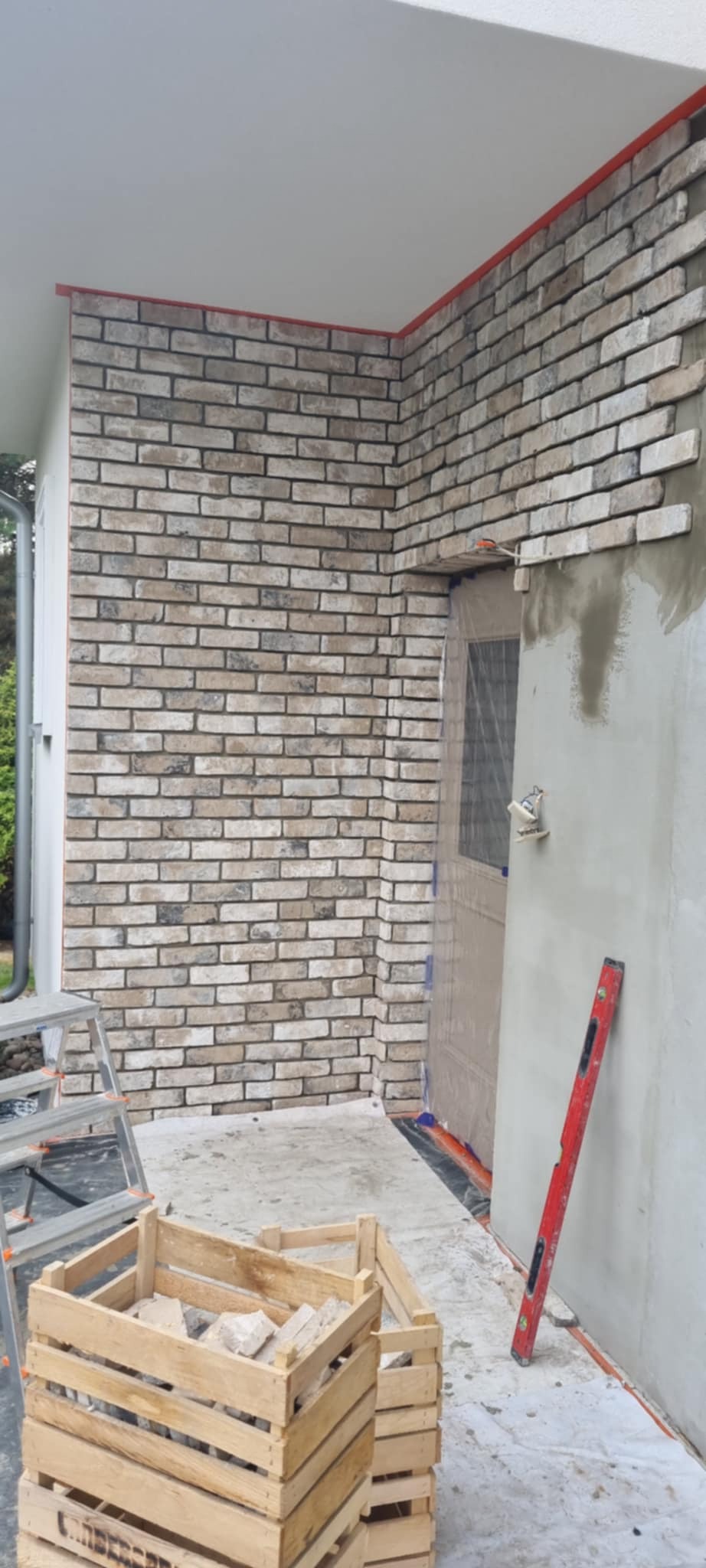 Kładzenie cegieł dekoracyjnych na ścianie zewnętrznej budynku. Widoczna skrzynka z cegłami, poziomica i drabina. Częściowo ukończona ściana z cegły.