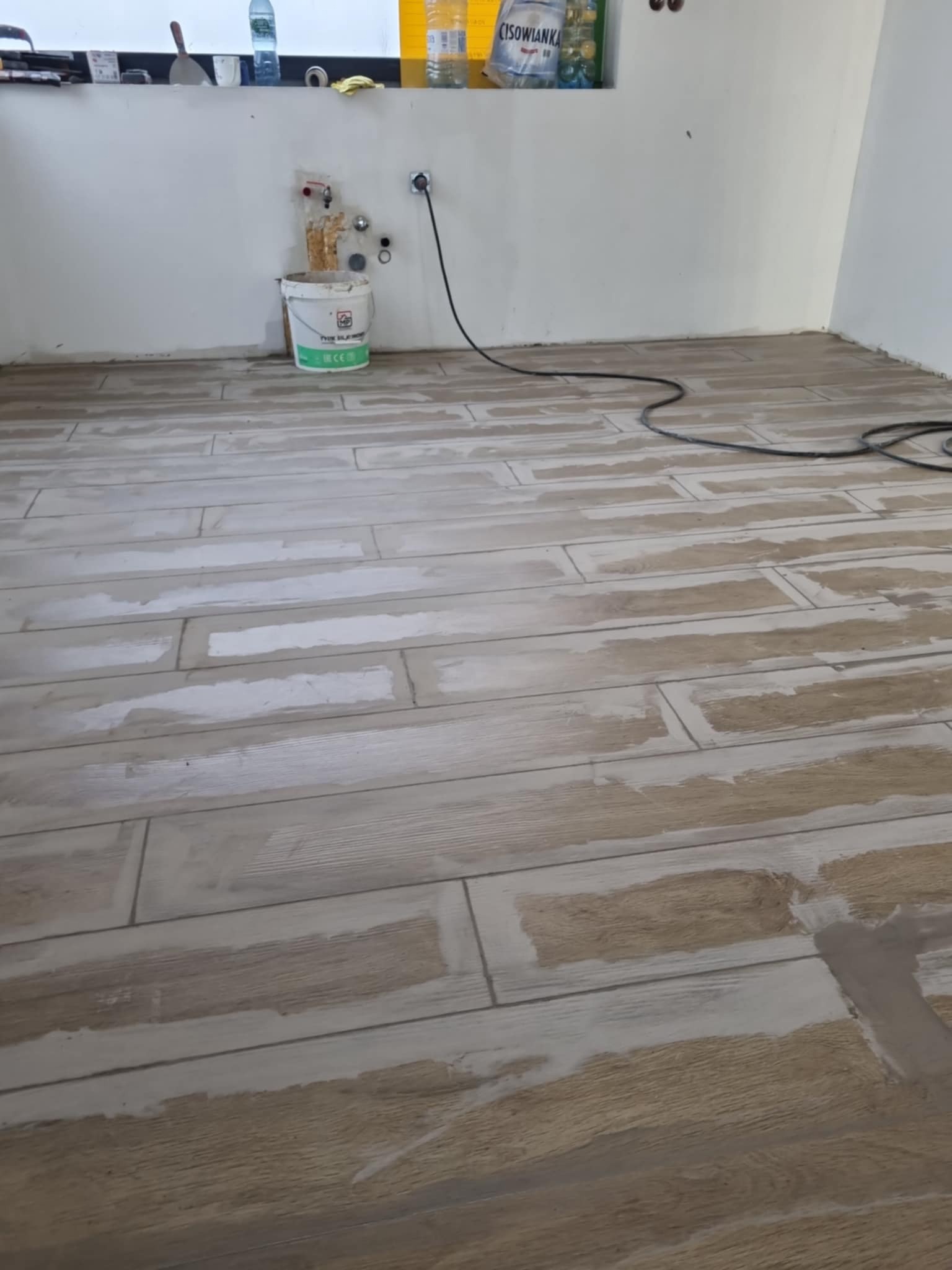 Ułożona podłoga z płytek imitujących drewno, z widocznymi fugami w trakcie wypełniania. W tle ściana z instalacją wodną i wiadro z zaprawą.