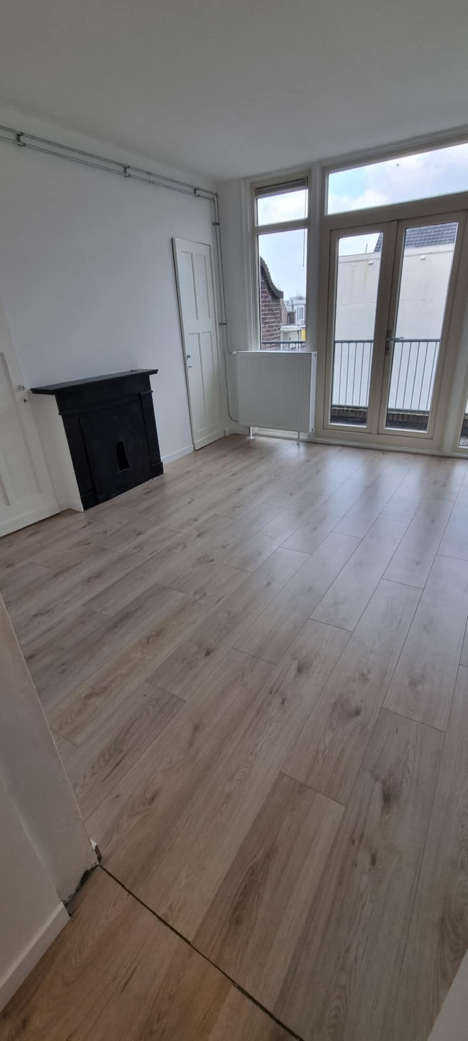 Jasne panele podłogowe w pokoju z białymi ścianami, czarnym kominkiem i balkonem. Widoczne drzwi i grzejnik. Neutralna kolorystyka, minimalistyczny wystrój.