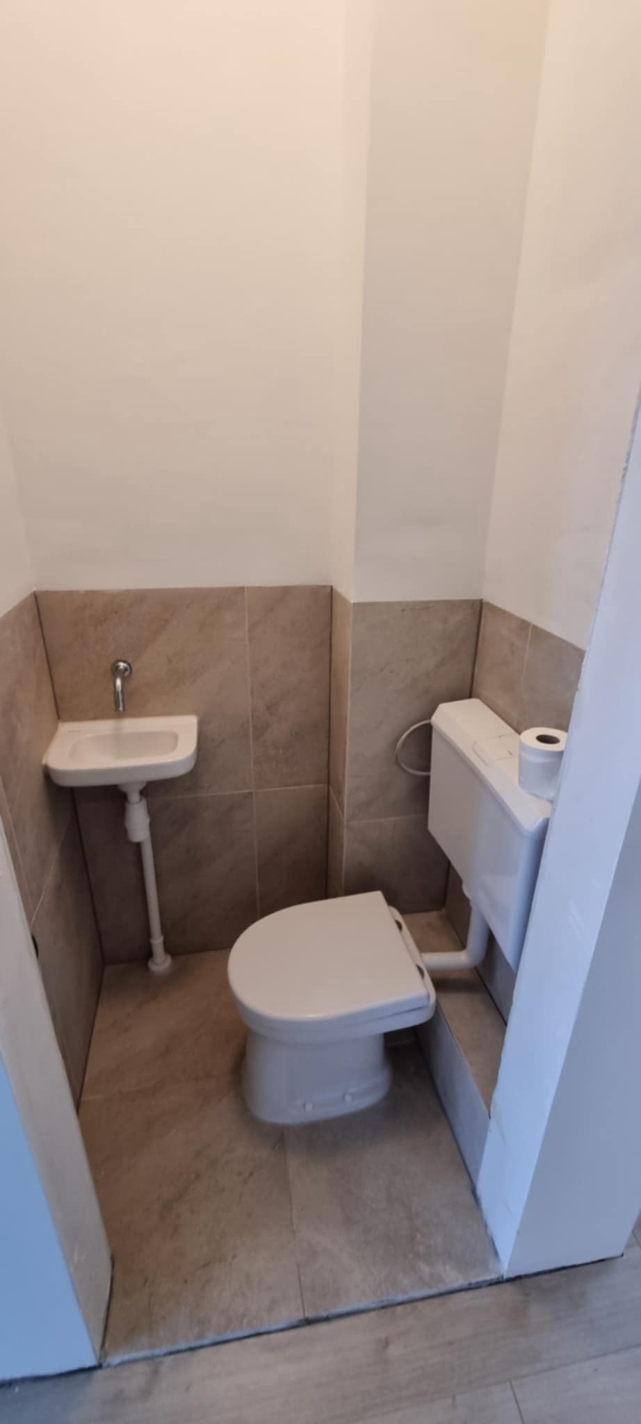 Mała łazienka po remoncie: biała toaleta i umywalka narożna na tle beżowych płytek. Widoczny fragment drzwi. Minimalistyczny design, czystość i funkcjonalność.